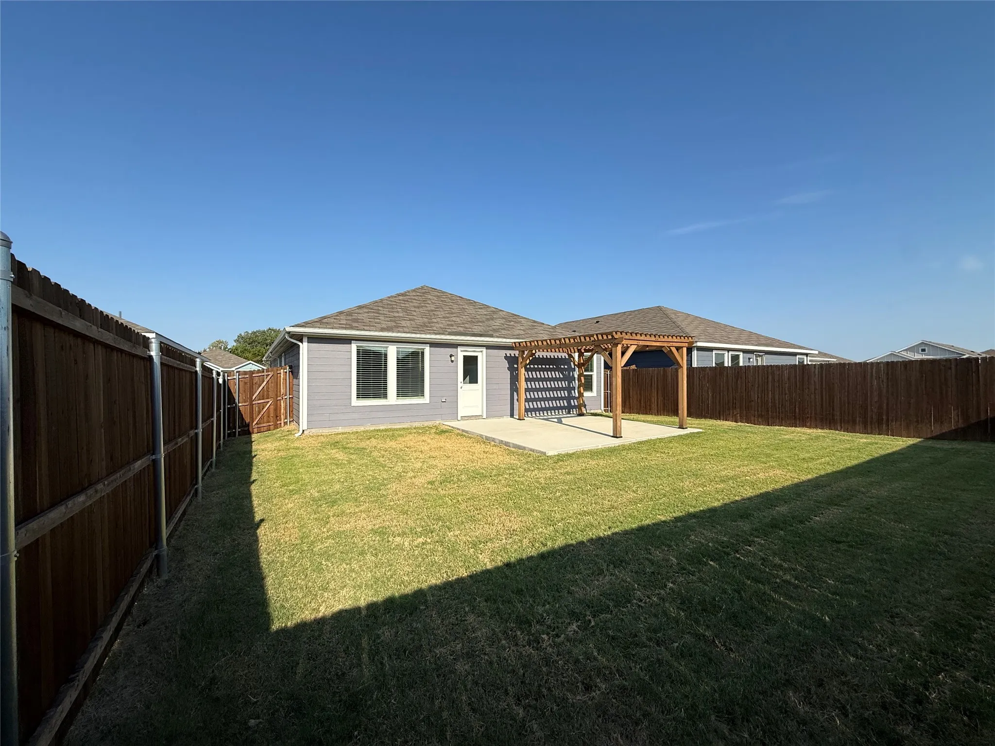 Expansive grass lawn with a concrete patio, wooden pergola, and privacy fencing