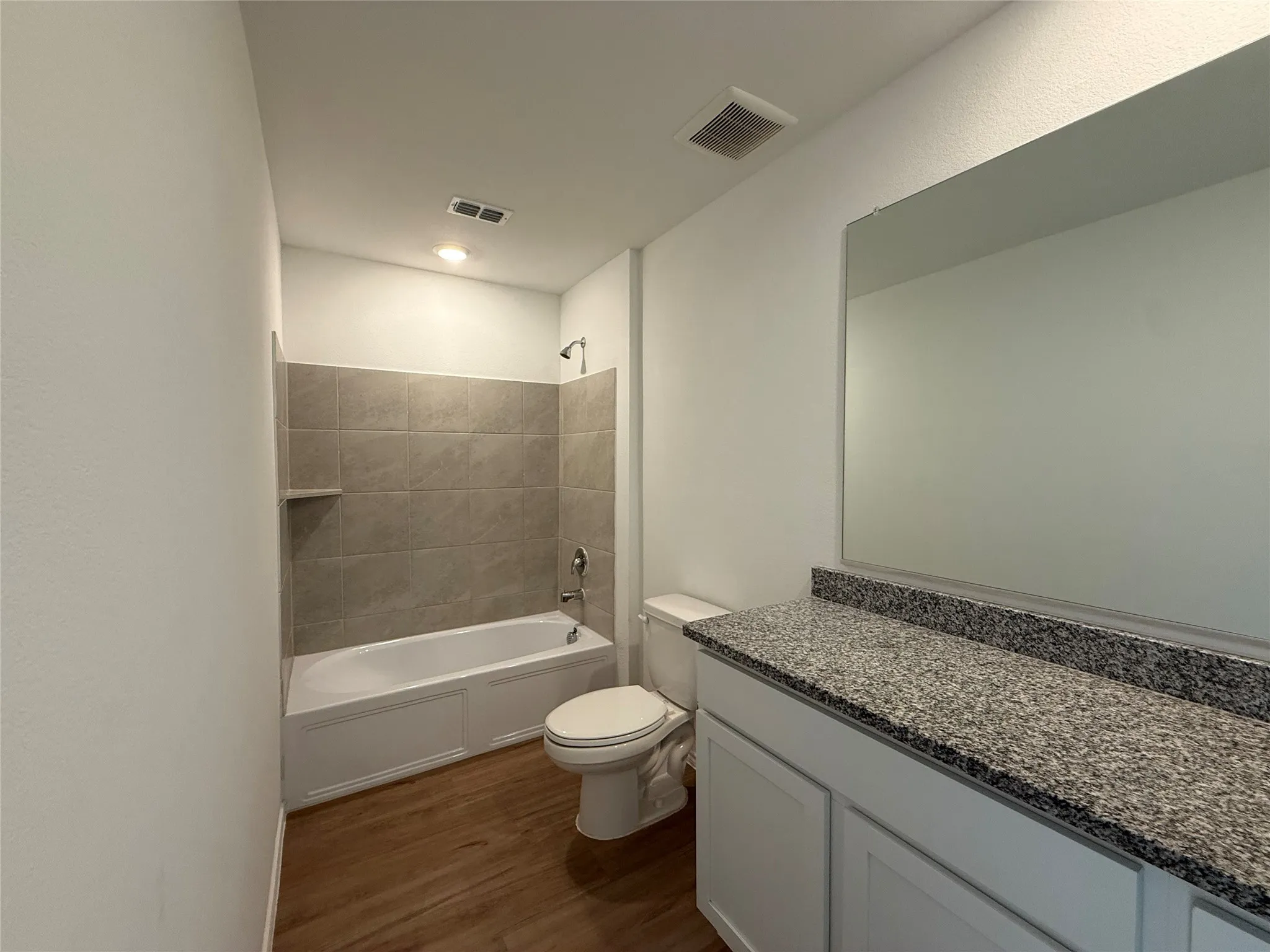 Wood-finish flooring, white vanity with dual cabinet doors, speckled countertop, frameless mirror, and tiled bath/shower combination