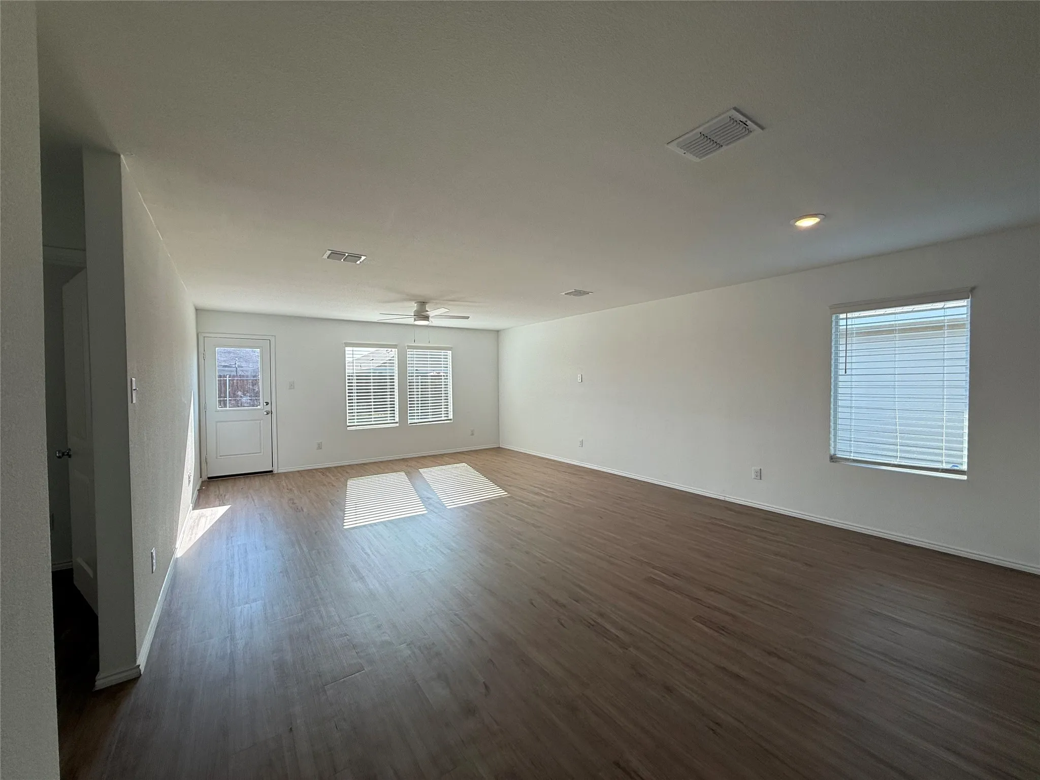 Spacious room featuring wood-finish flooring, white walls, multiple windows with blinds, a white ceiling fan, and recessed lighting