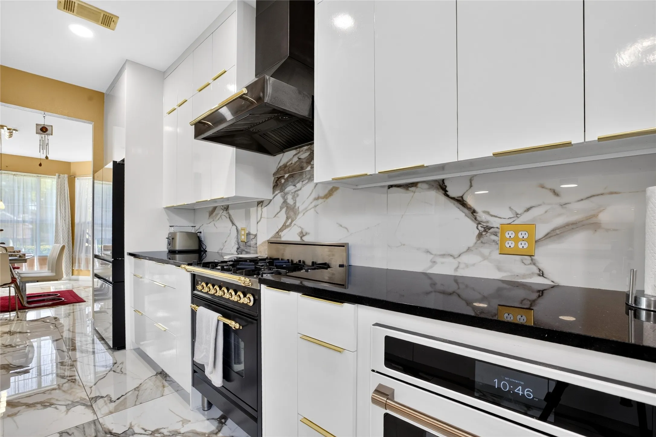Contemporary kitchen featuring glossy white cabinetry, black countertops, a marbled backsplash, a black range with gold-toned accents, and integrated appliances