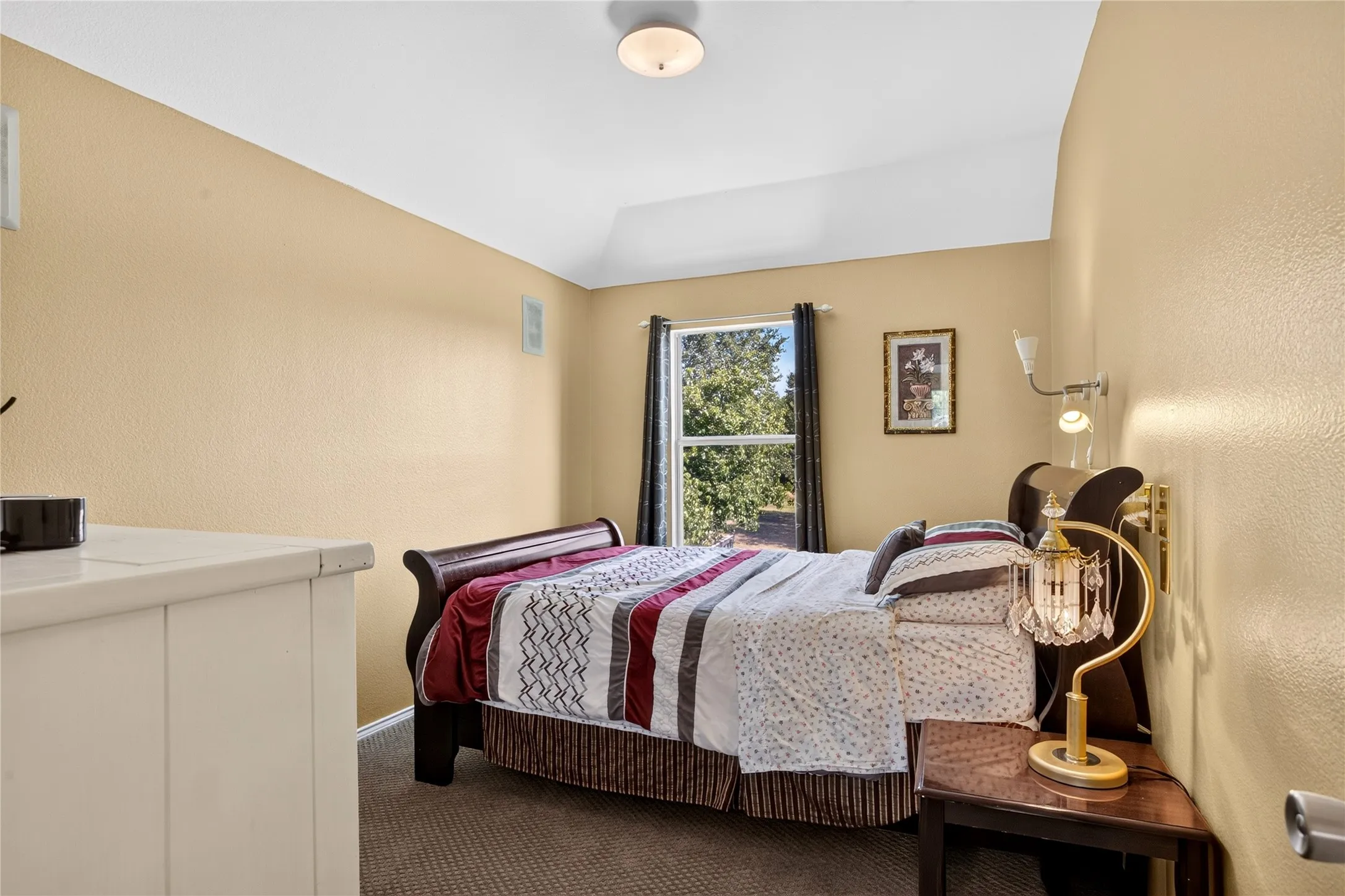 Vaulted ceiling, wall-mounted light fixture, window with drapes, carpeted flooring, and a window framing mature trees