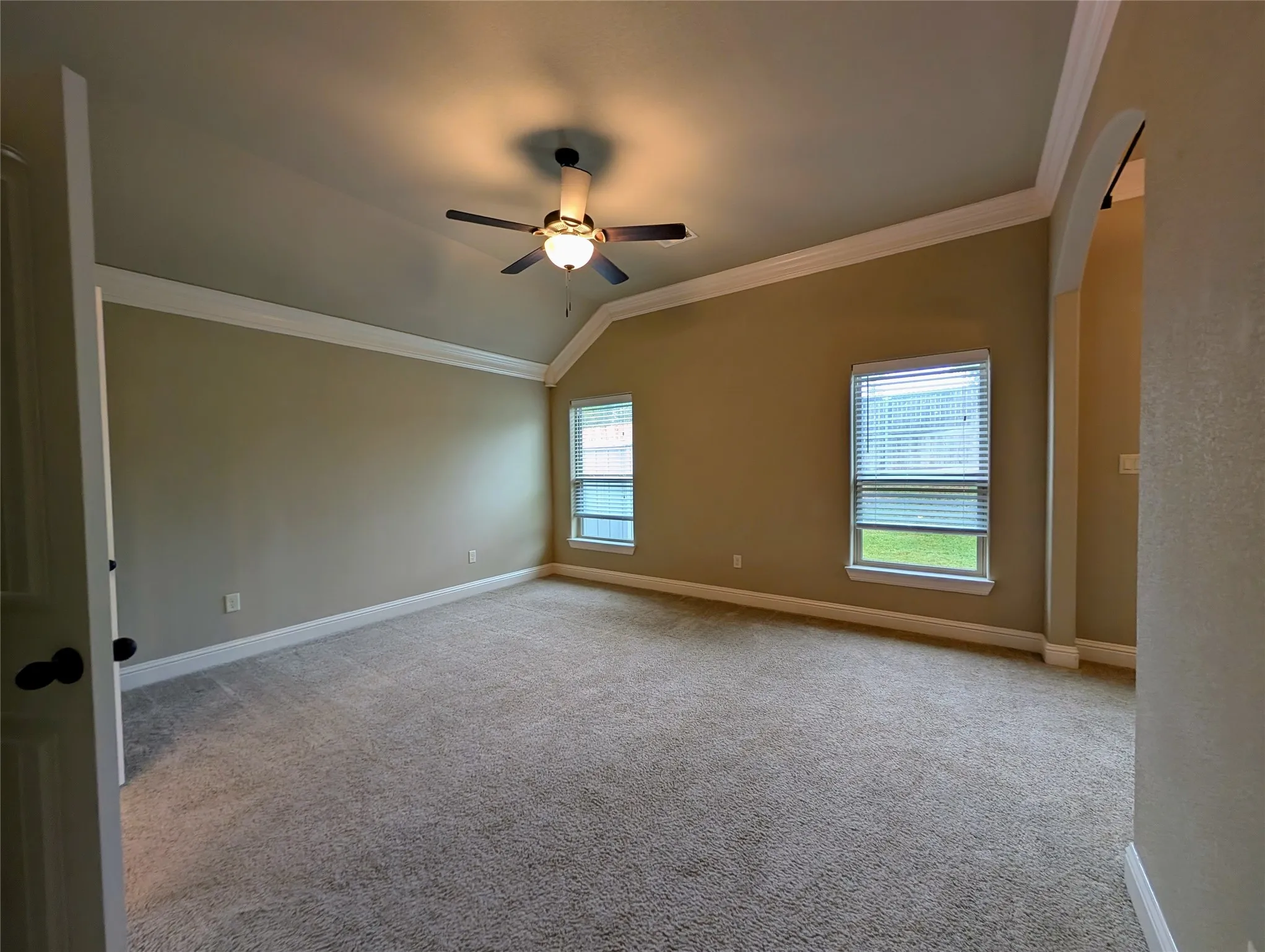Vaulted ceiling, ceiling fan with integrated lighting, two windows with blinds, neutral-toned carpeting, and a broad arched entryway
