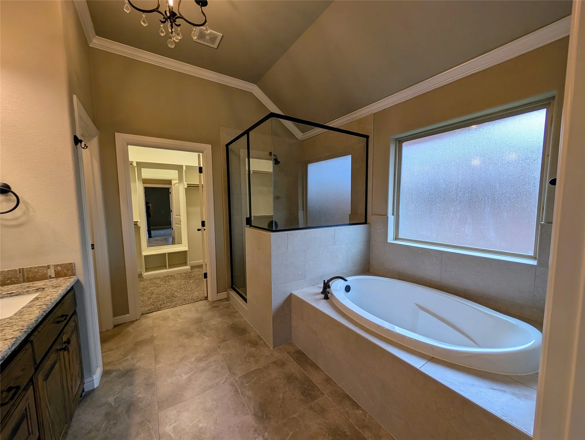Oversized soaking tub, separate glass-enclosed shower, wood-finish vanity with stone countertop, tile flooring, and vaulted ceiling