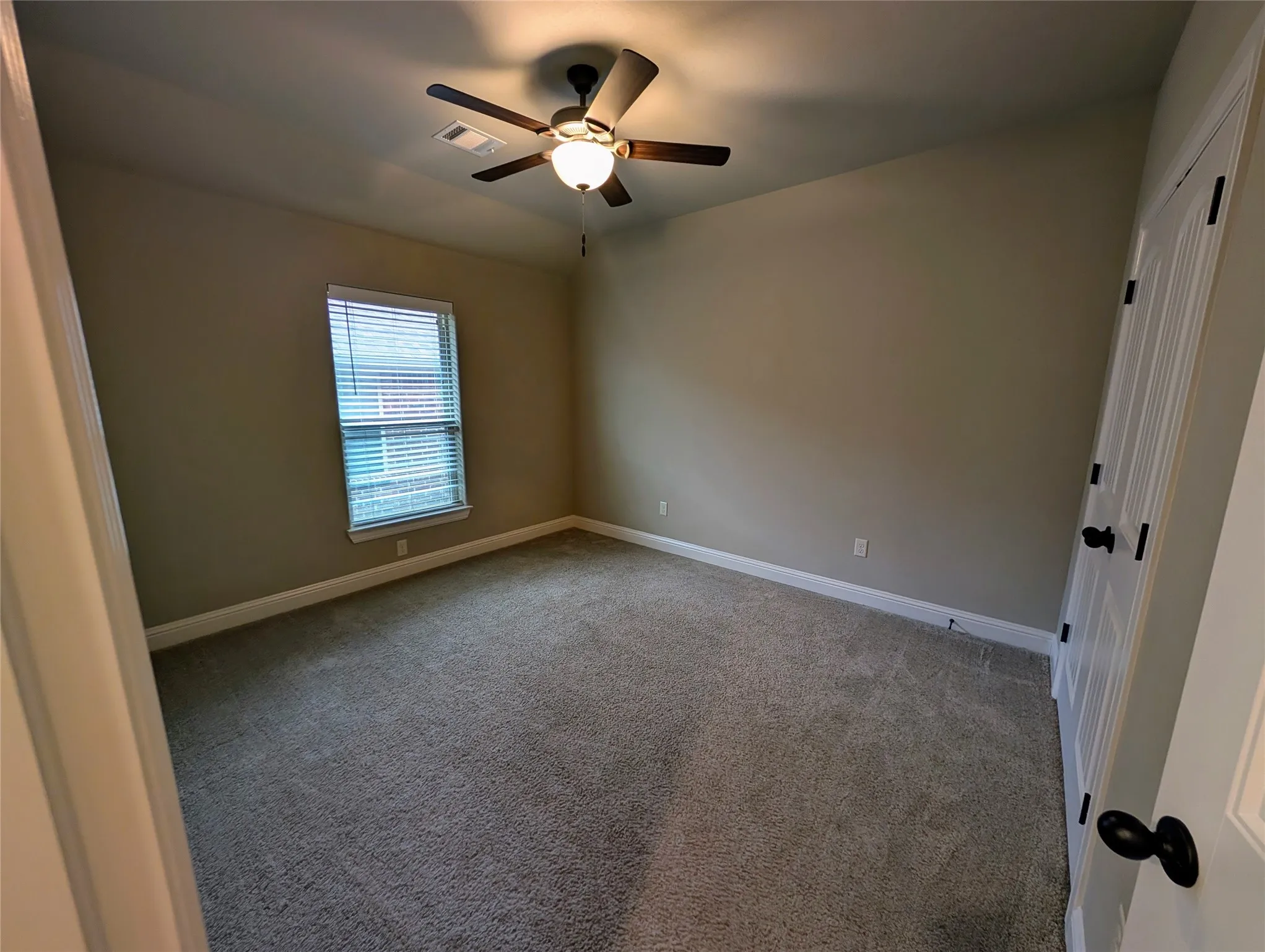 Neutral wall paint, plush carpeting, ceiling fan with integrated lighting, window with blinds, and a paneled door with dark hardware