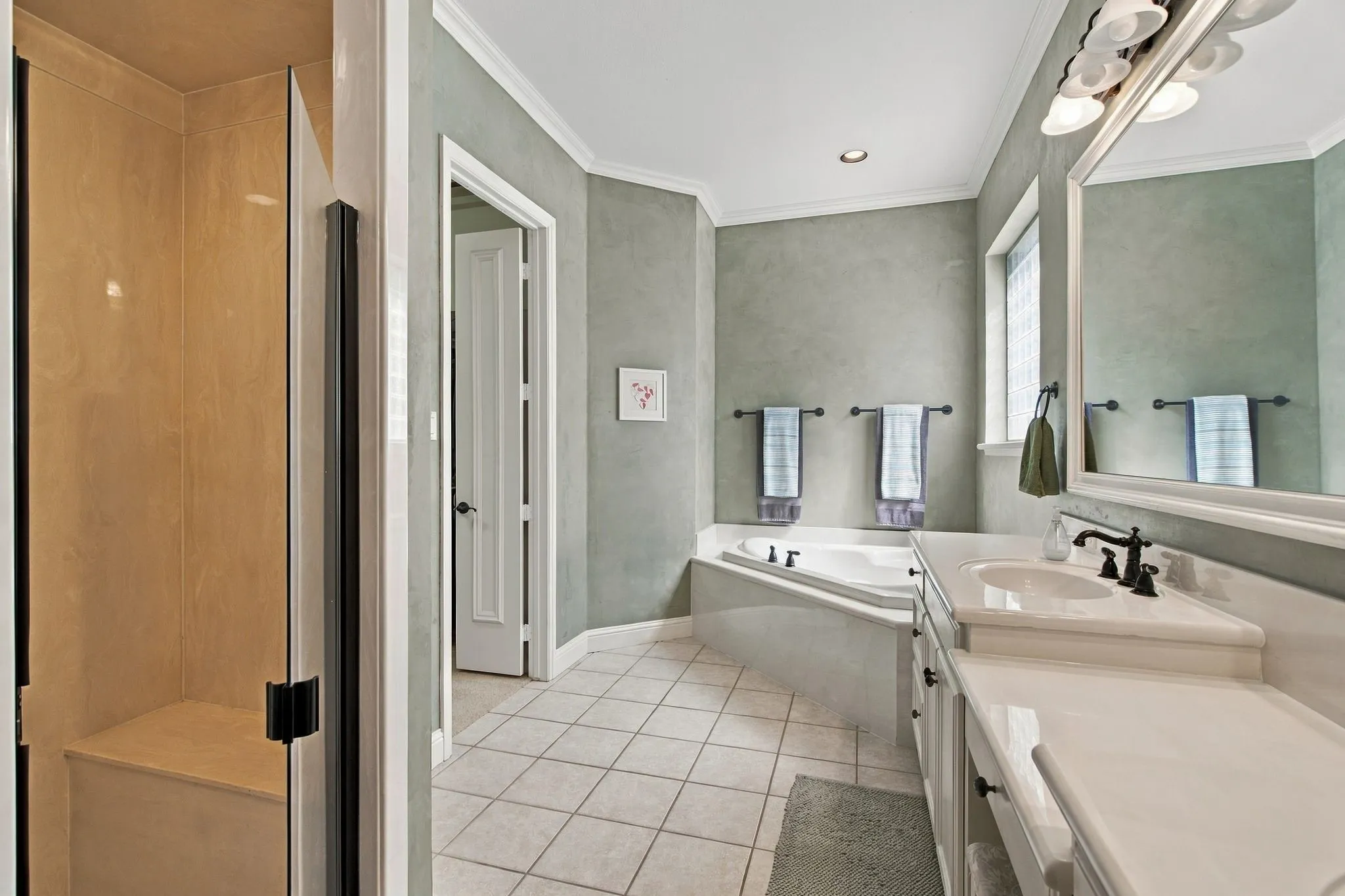 White tile flooring, built-in soaking tub, vanity with integrated sink, rectangular mirror, and an enclosed shower with a built-in bench
