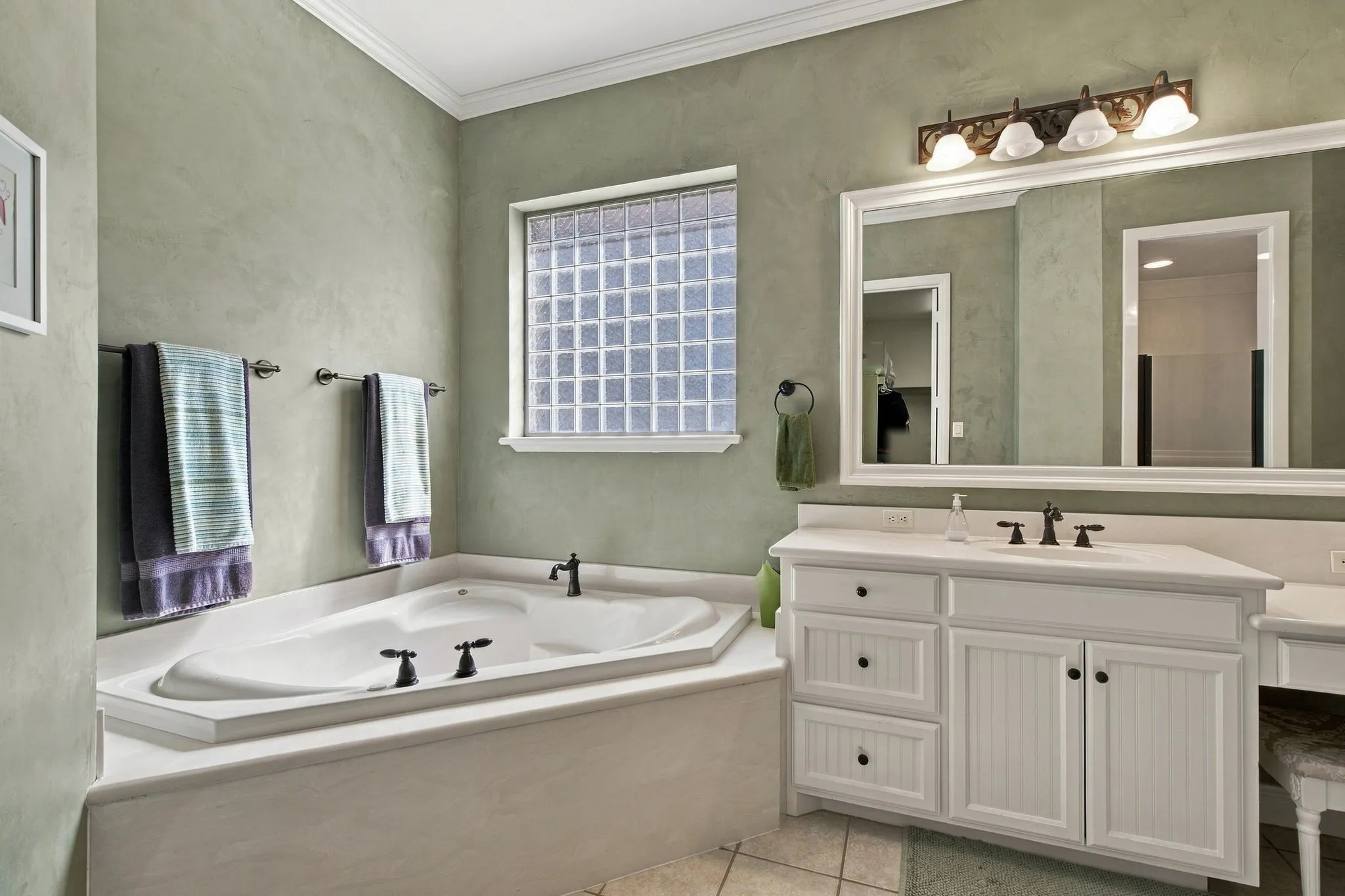 Built-in soaking tub with dark bronze fixtures, glass block window, white vanity with beadboard paneling, integrated sink, and large framed mirror