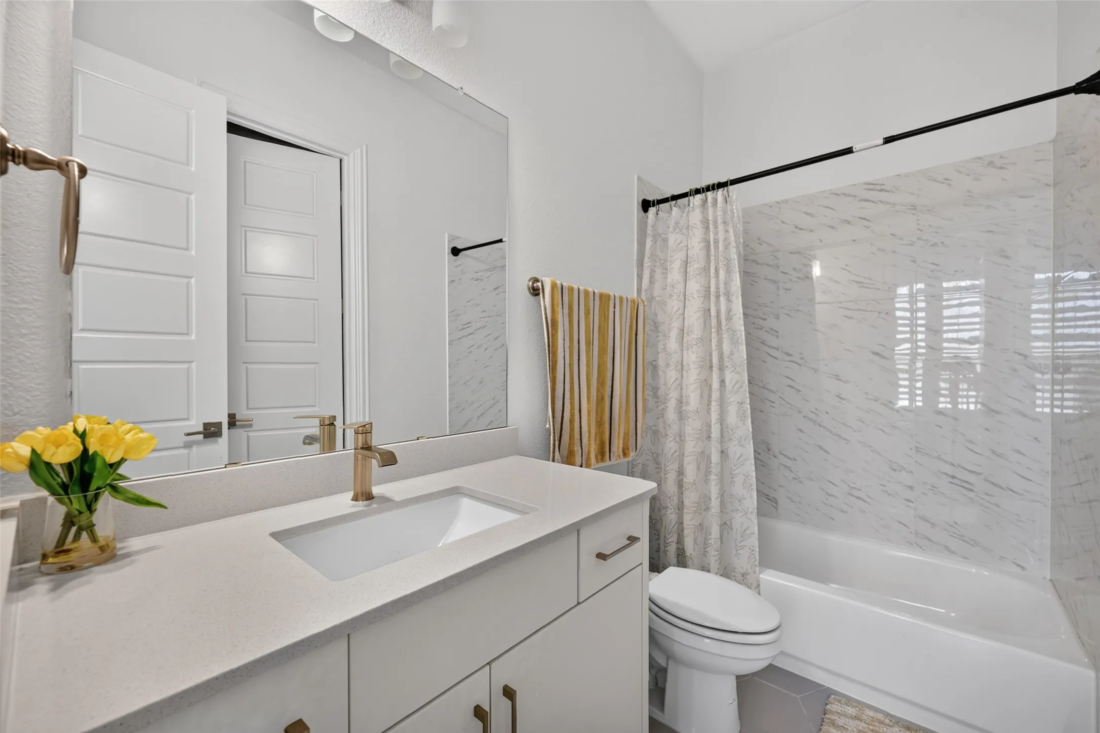 Vanity with integrated sink and brass-finish faucet, quartz countertop, white cabinetry with brass-finish hardware, and a tiled shower/tub combination with black-finish fixtures
