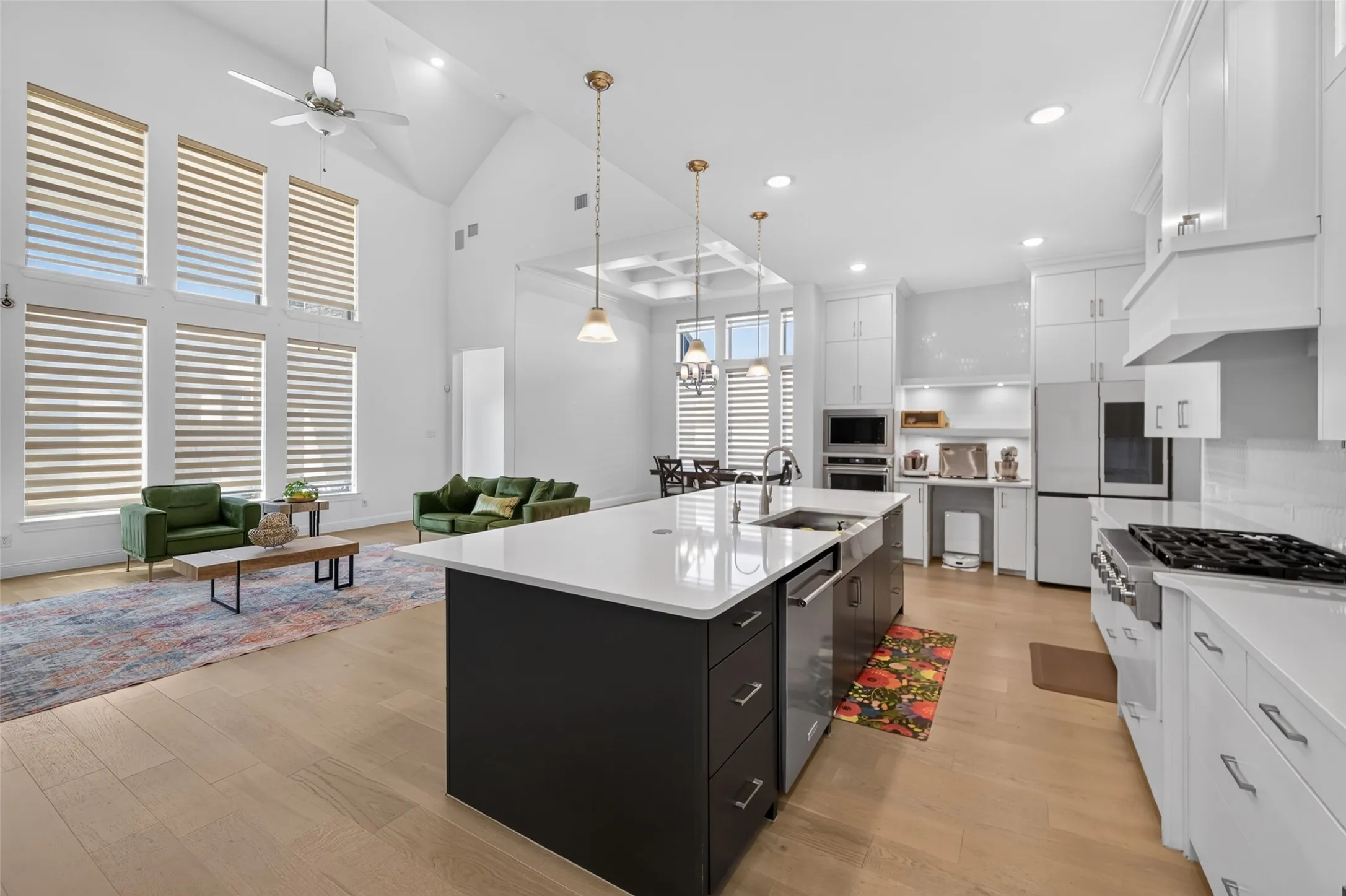 Open-concept living space featuring vaulted ceilings, wood-finish flooring, a kitchen island with an integrated sink, white cabinetry, and stainless steel appliances