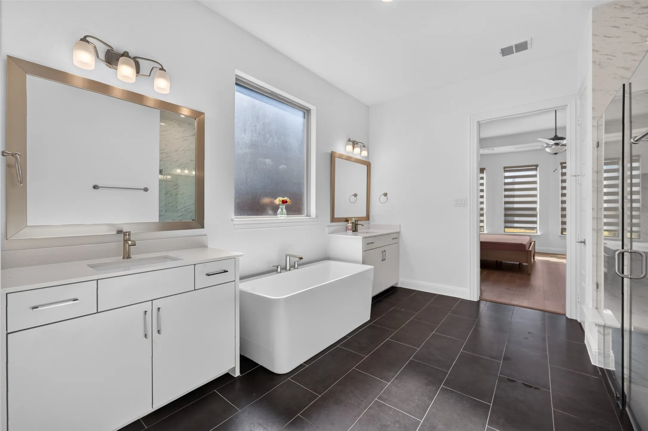 Bathroom featuring dual vanities with integrated sinks, a freestanding soaking tub, dark tile flooring, and a glass-enclosed shower