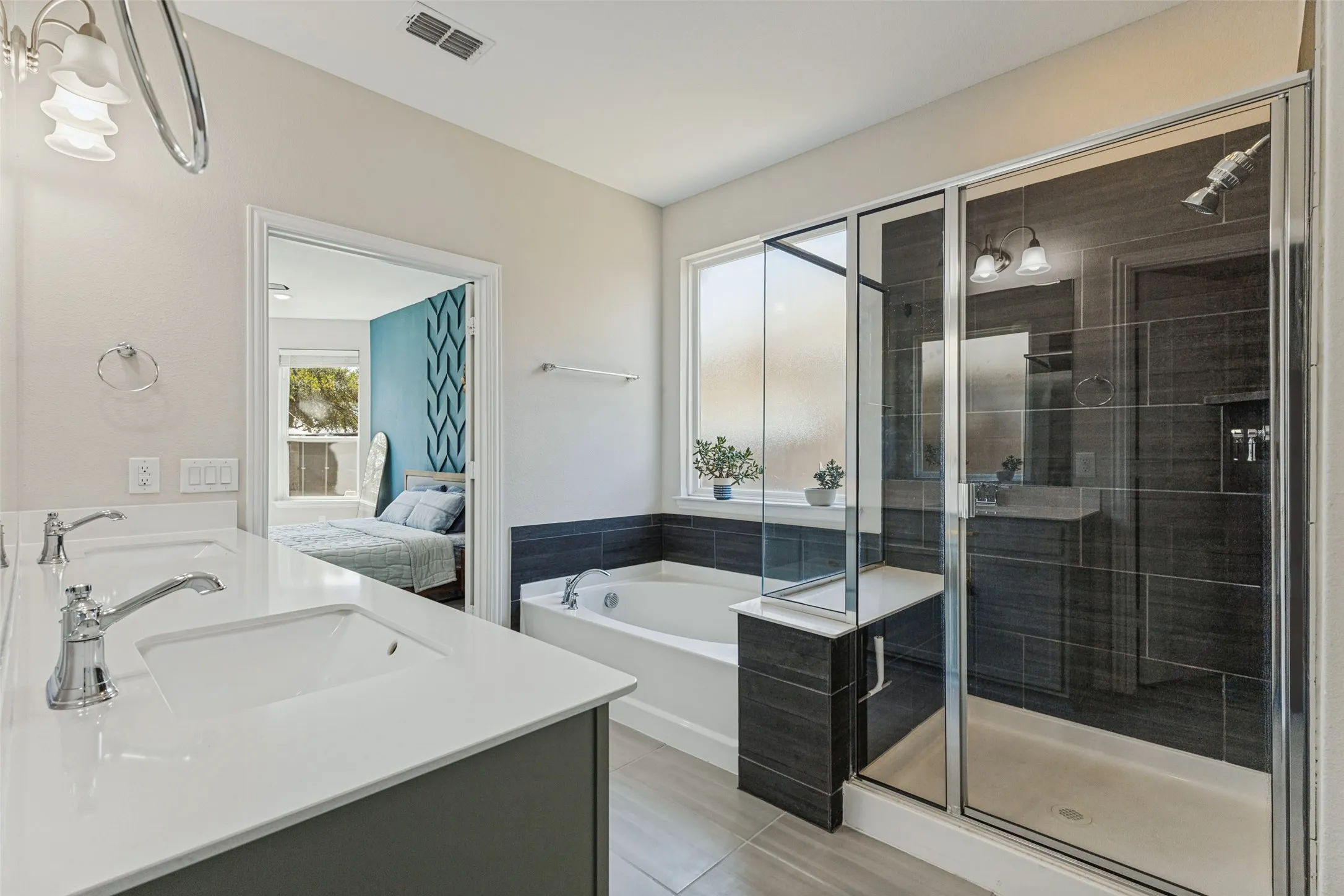 Double vanity with integrated sinks and chrome fixtures, large soaking tub, glass-enclosed walk-in shower with dark tile surround, and frosted window
