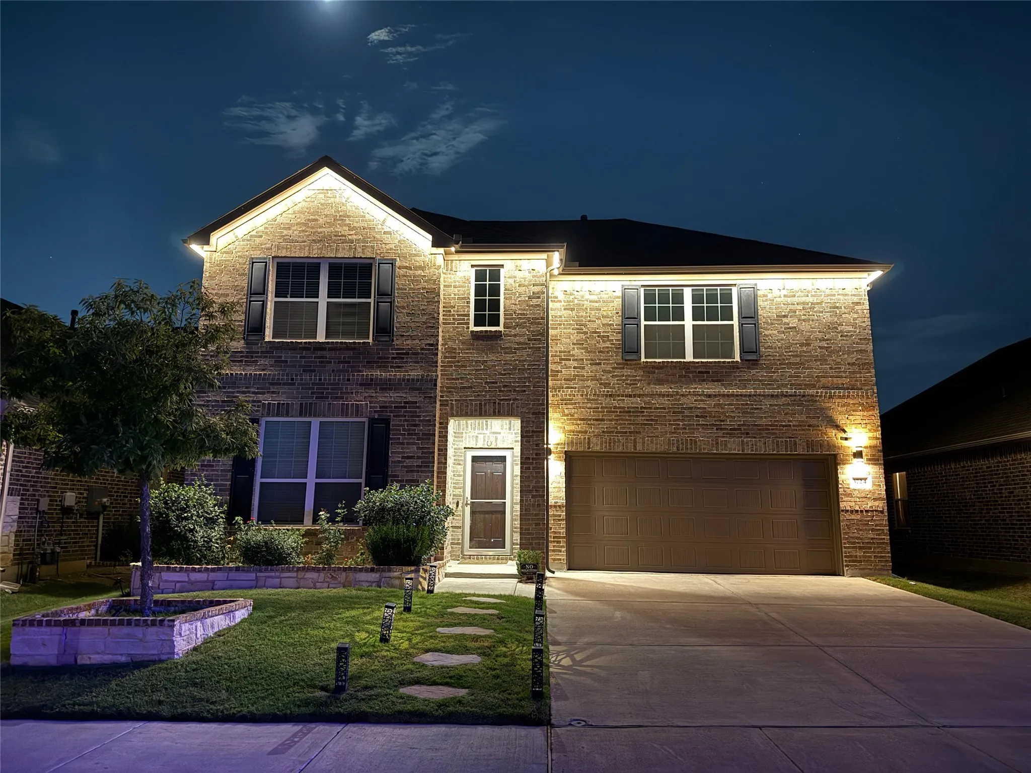 Brick facade with illuminated accent lighting, a two-car garage, and a landscaped front yard featuring a stone-edged planter and stepping stones