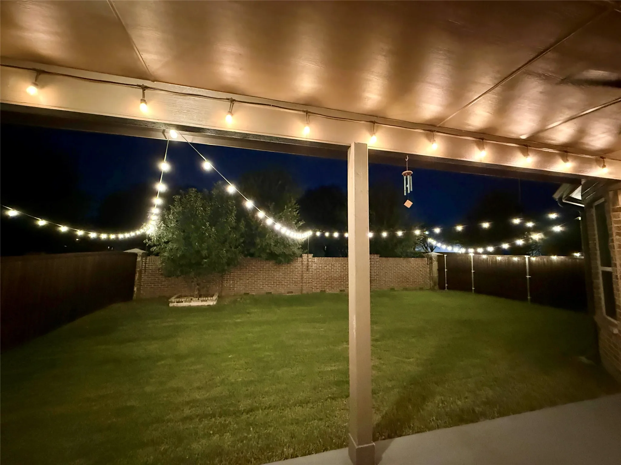 Covered patio with string lighting, expansive lawn, mature tree, and brick fencing