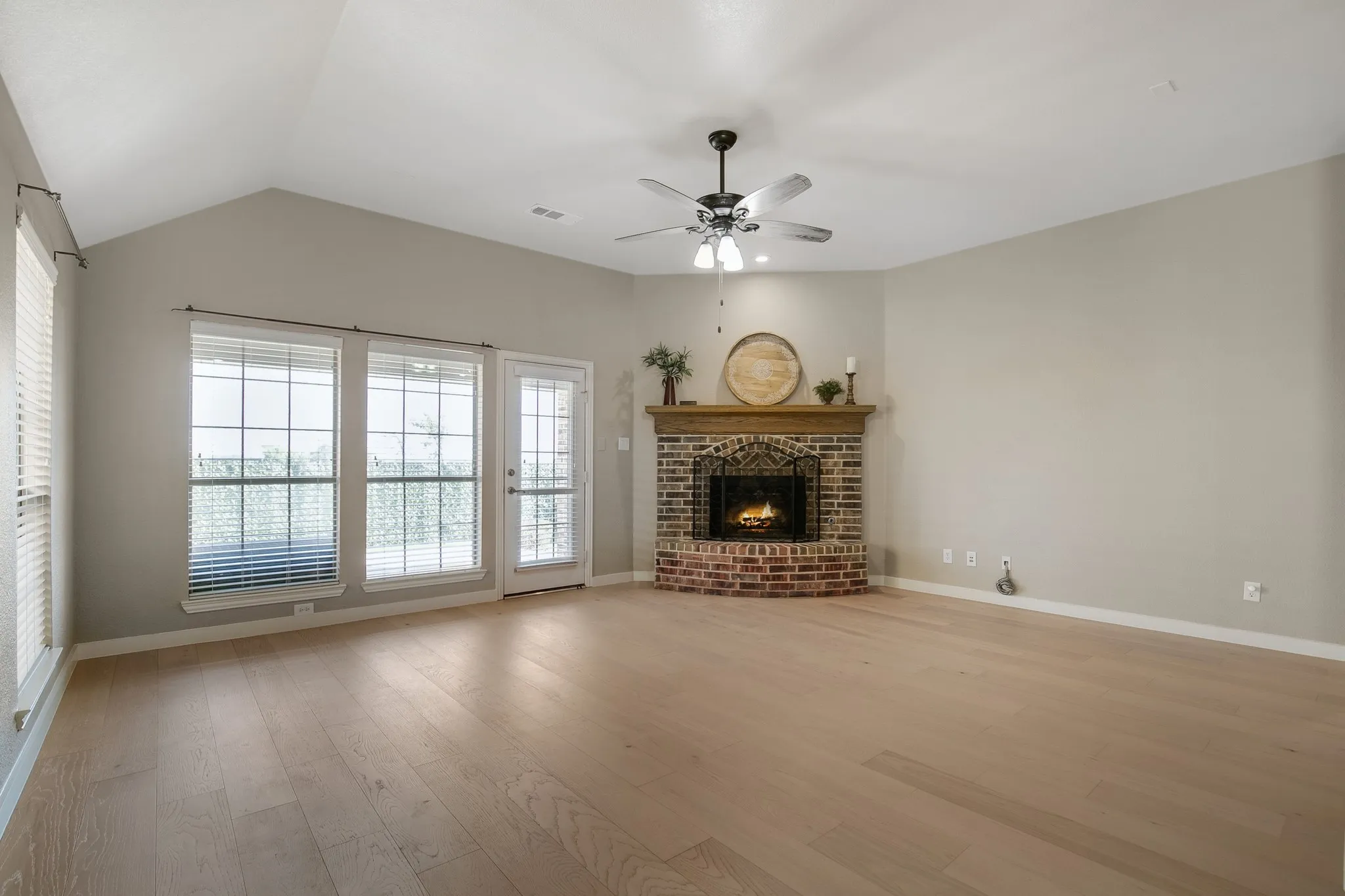 Vaulted ceiling, corner brick fireplace with wood mantel, ceiling fan, wood-finish flooring, and multiple windows