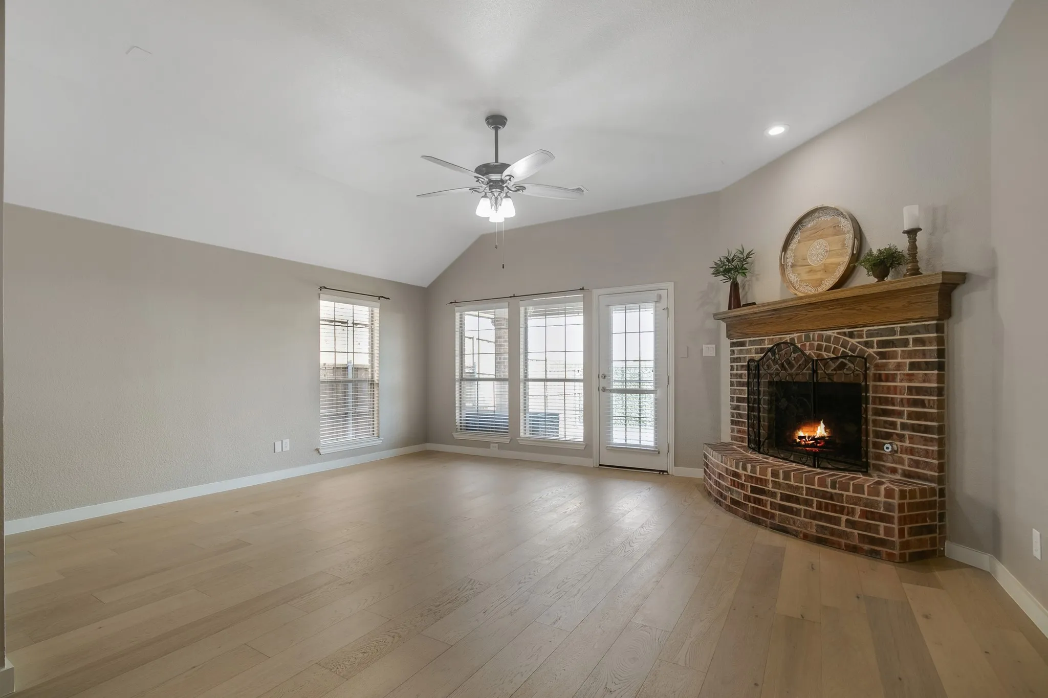 Vaulted ceilings with a ceiling fan, light wood-finish flooring, brick fireplace with a wood mantel, and multiple windows