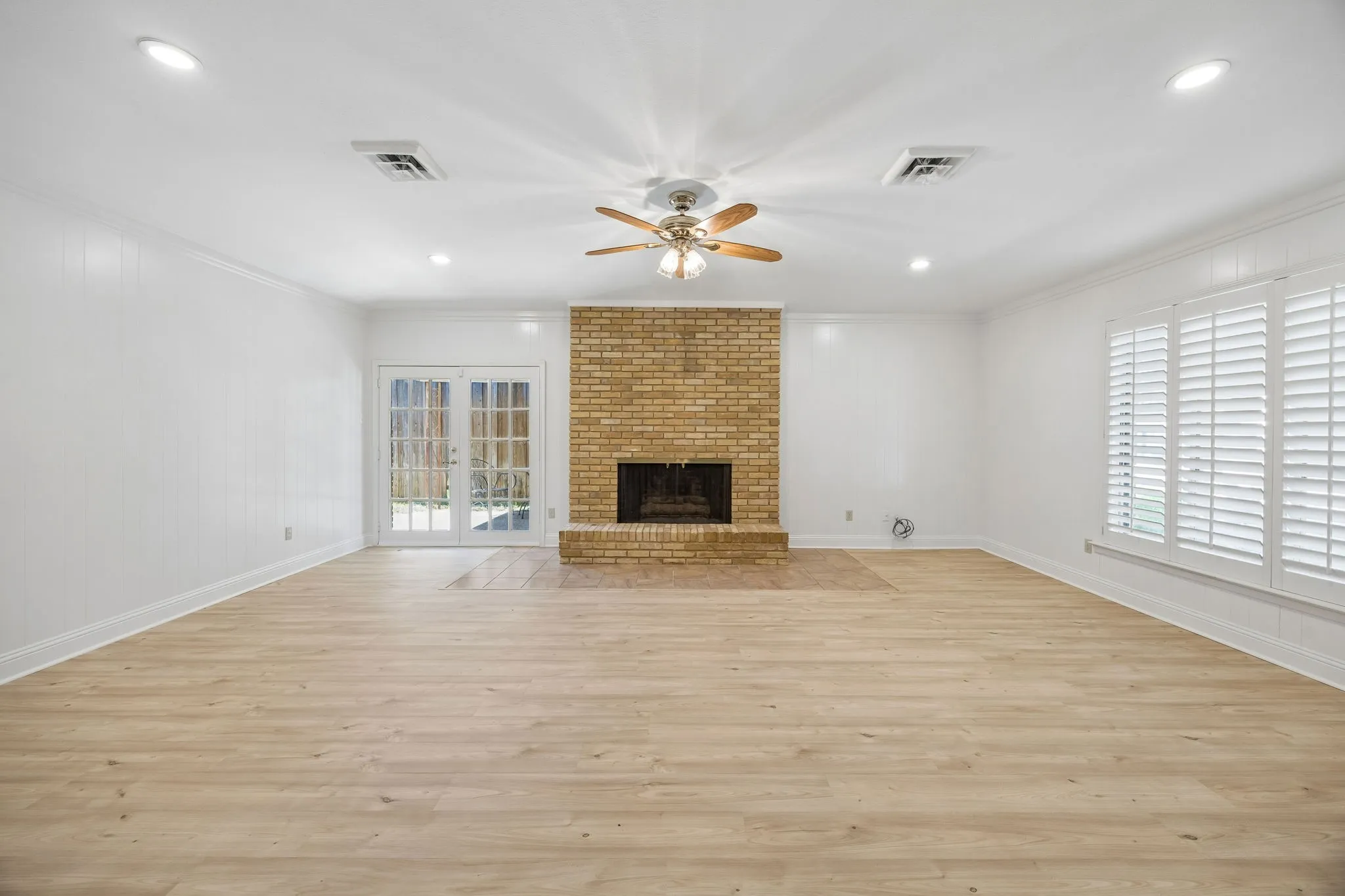 Spacious living room with a brick fireplace, wood-look flooring, plantation shutters, and French doors to the backyard.