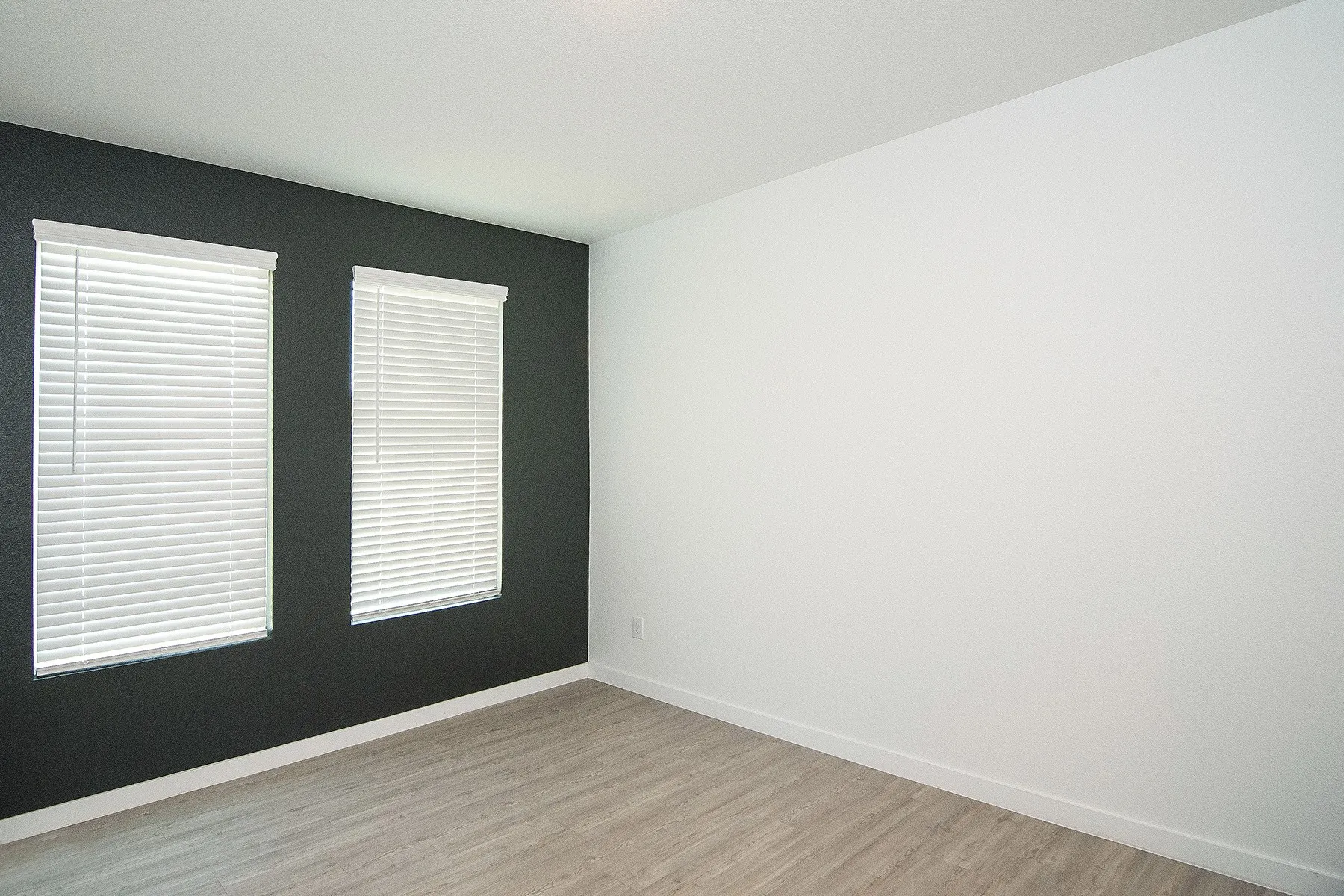 Dual windows with white blinds, dark accent wall, light wood-finish flooring, and white walls