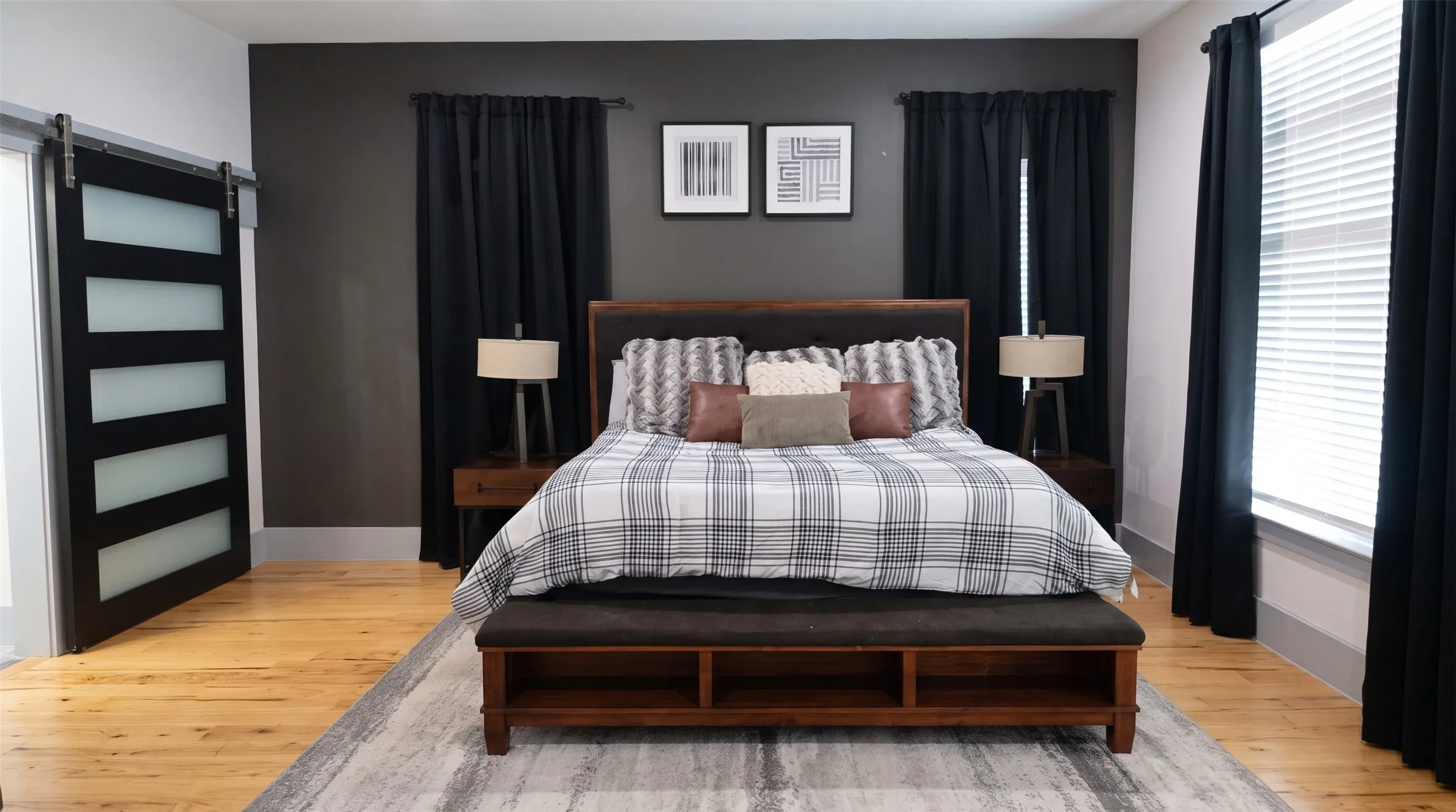 Hardwood flooring, sliding barn door with frosted glass panels, dark accent wall, window with blinds and dark curtains, and a wood-frame bed with an upholstered headboard
