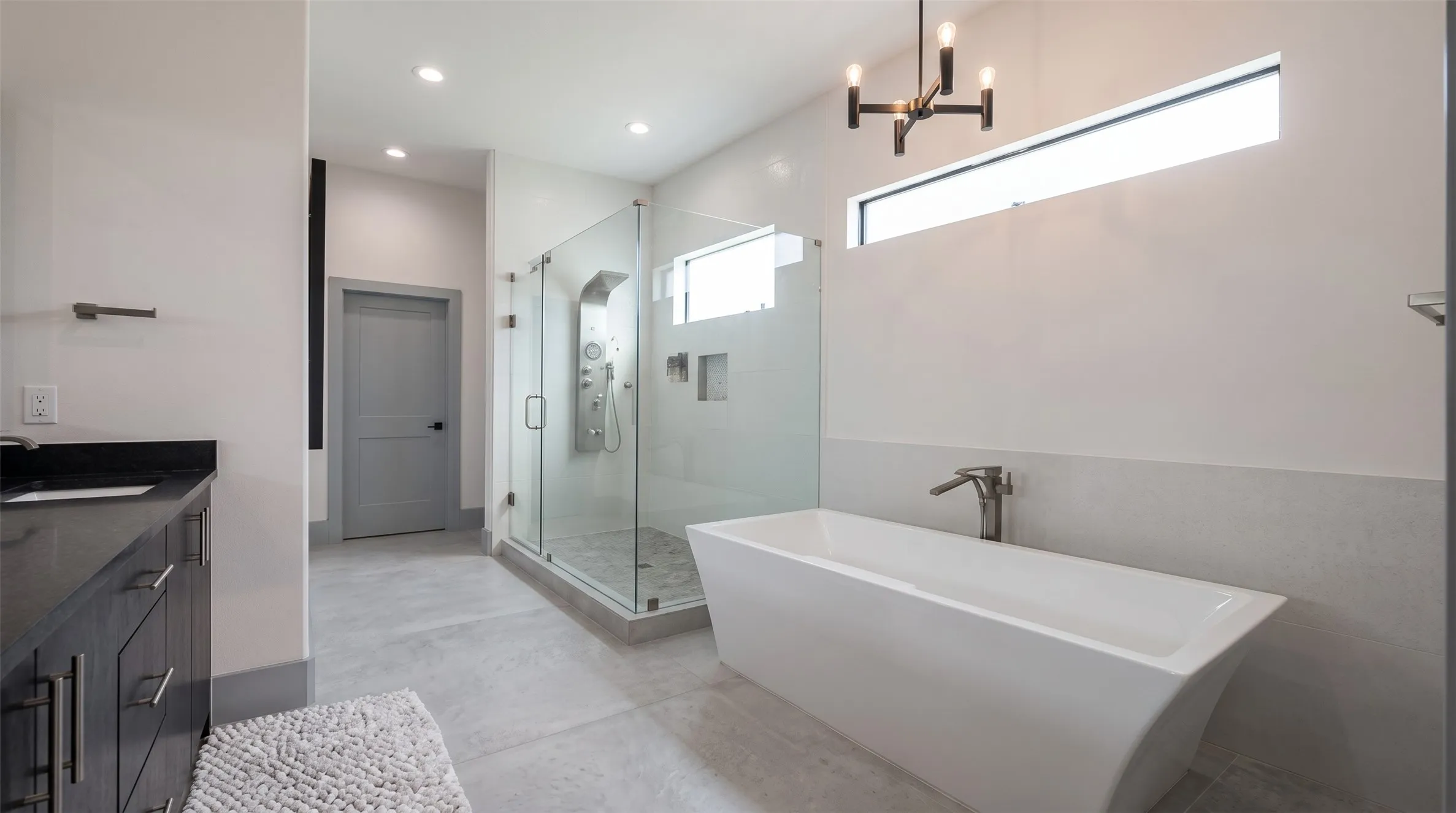 Freestanding soaking tub with a modern faucet, glass-enclosed shower with multiple showerheads, dark wood-finish vanity with a dark countertop, and a contemporary chandelier