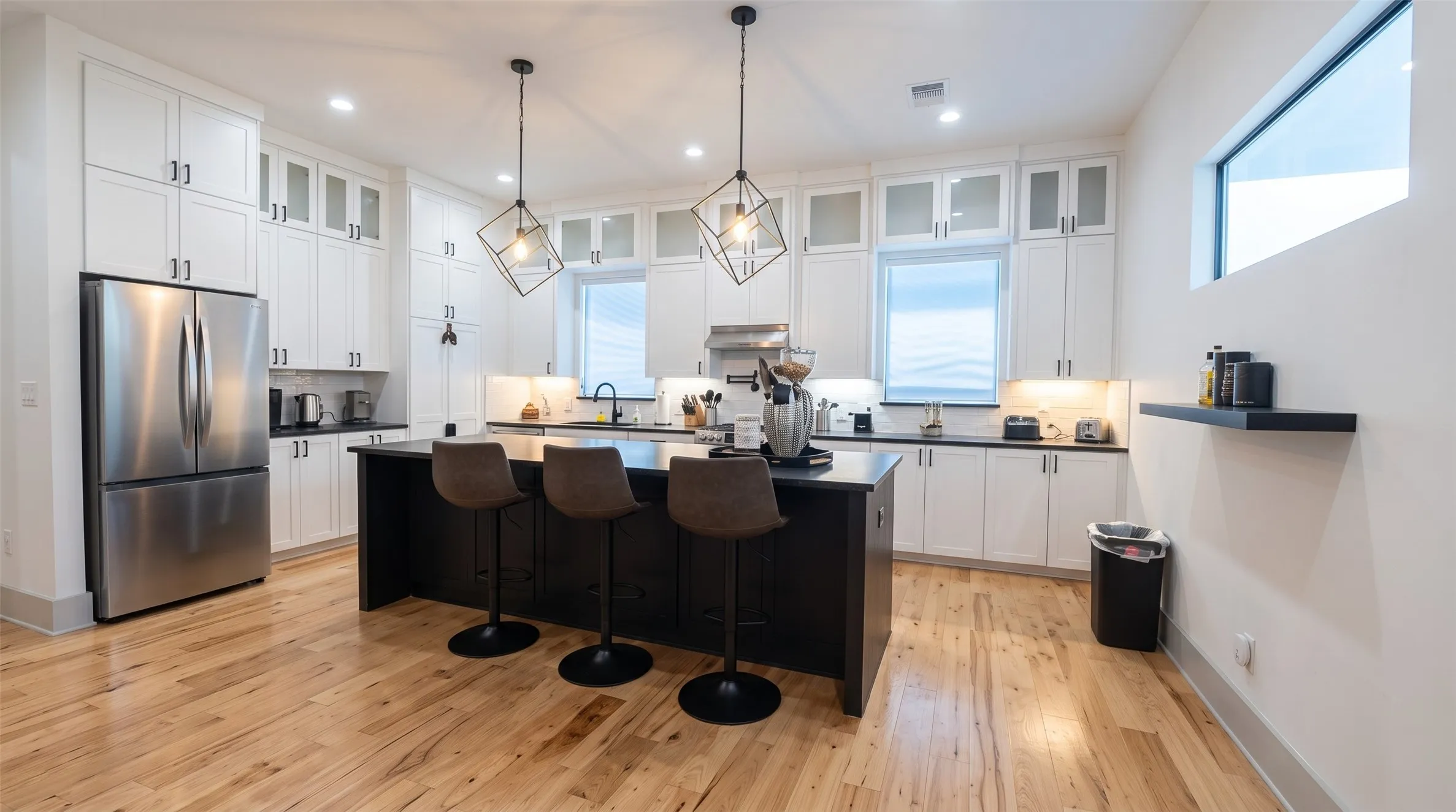 Modern kitchen featuring a dark island with seating, stainless steel appliances, white cabinetry with dark hardware, and light wood-finish​​‌​​​​‌​​‌‌​‌‌​​​‌‌​‌​‌​‌​​​‌​​ flooring