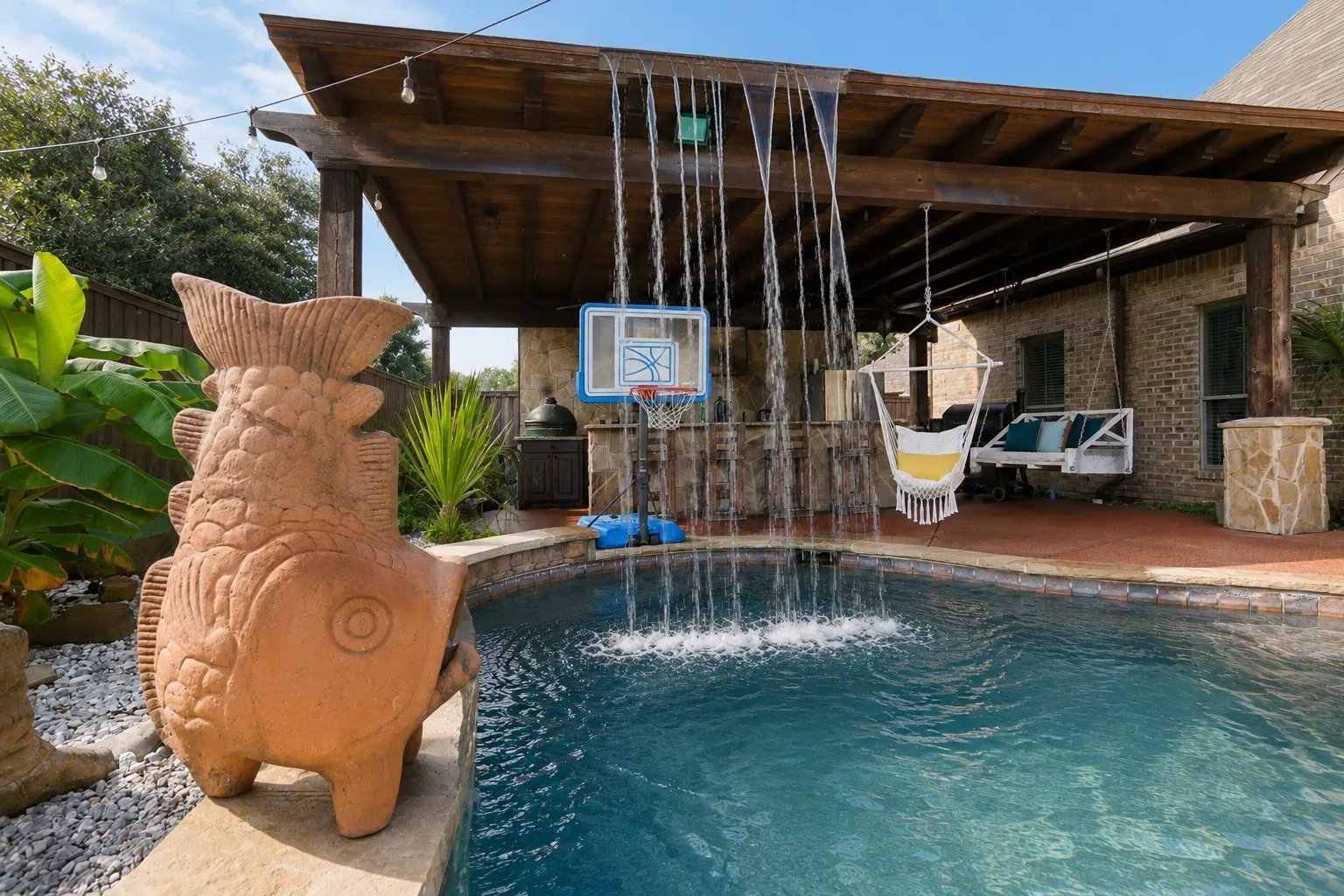 Swimming pool with water features, wood pergola, brick exterior, and a basketball hoop