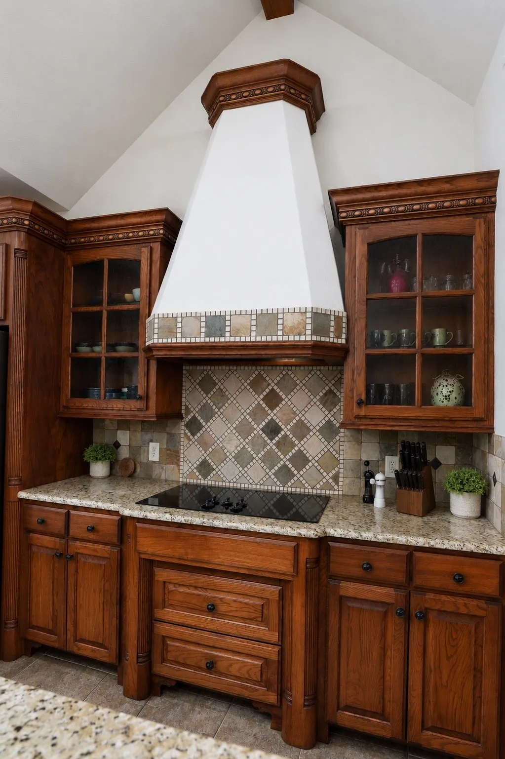 Vaulted ceiling, custom wood cabinetry, glass-front upper cabinets, patterned tile backsplash, and built-in cooktop