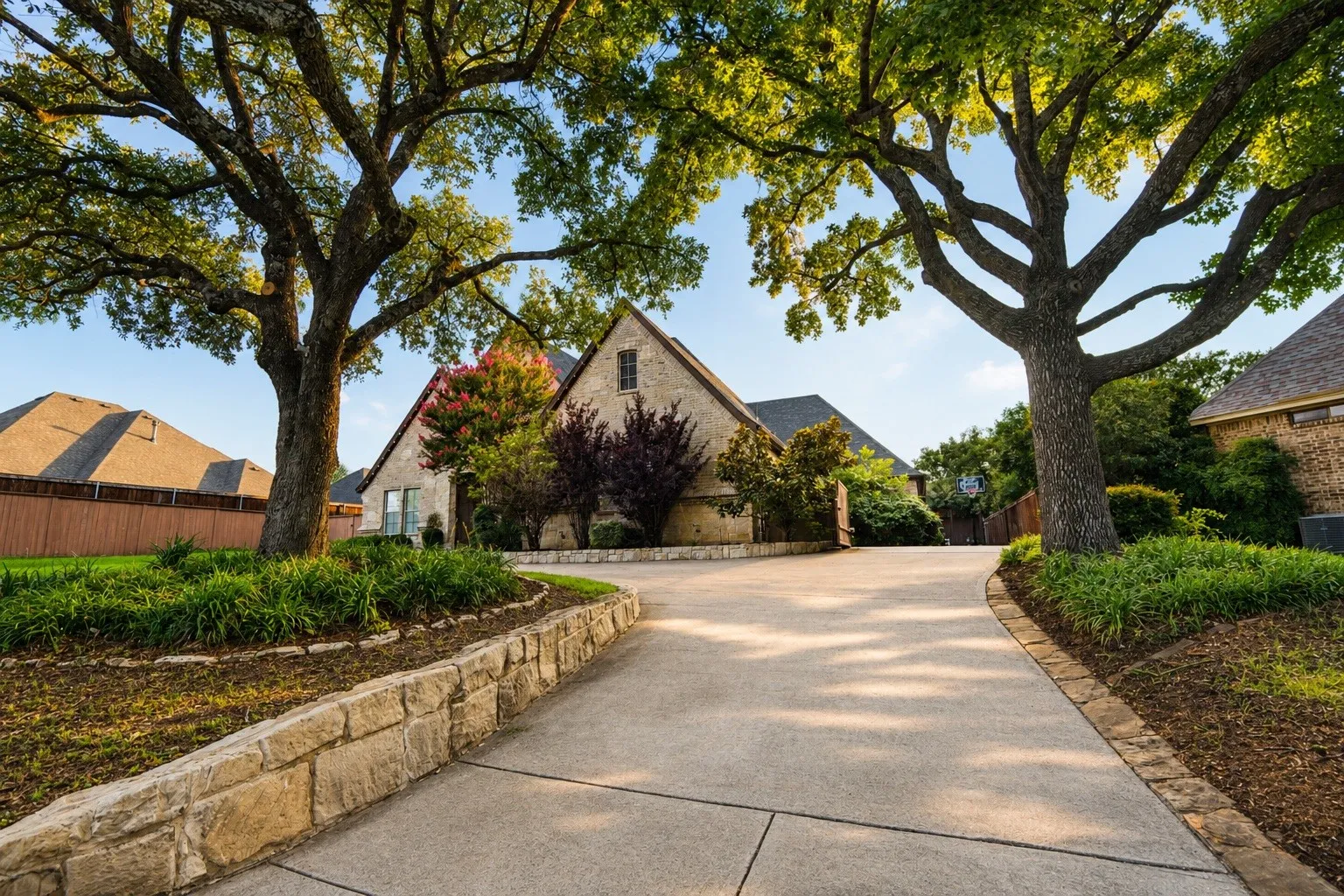 Stone facade residence with prominent gables, a concrete drive, and mature trees, complemented by stone retaining walls