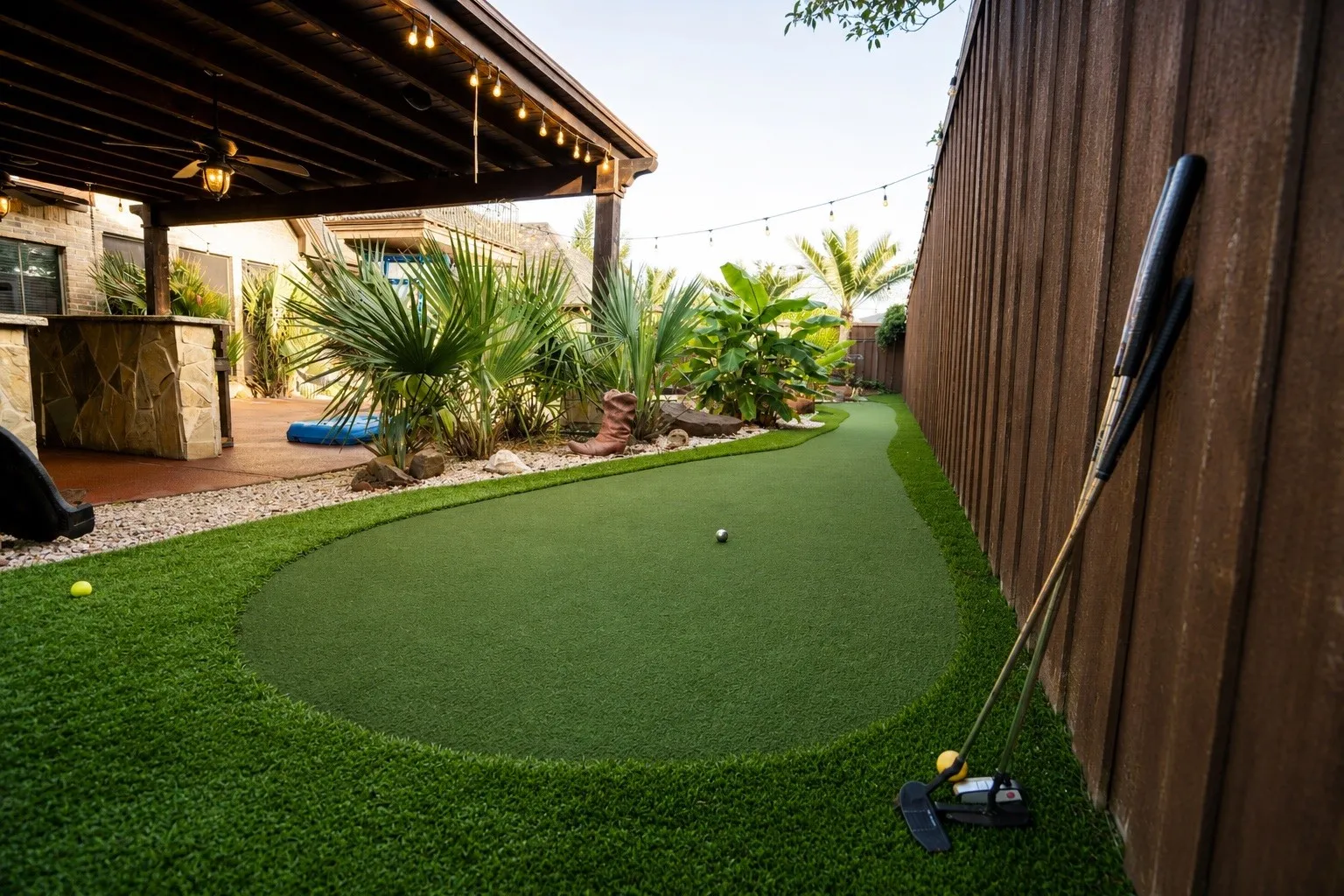 Backyard putting green with artificial turf, stone-clad outdoor bar, covered patio structure, mature landscaping, and privacy fencing