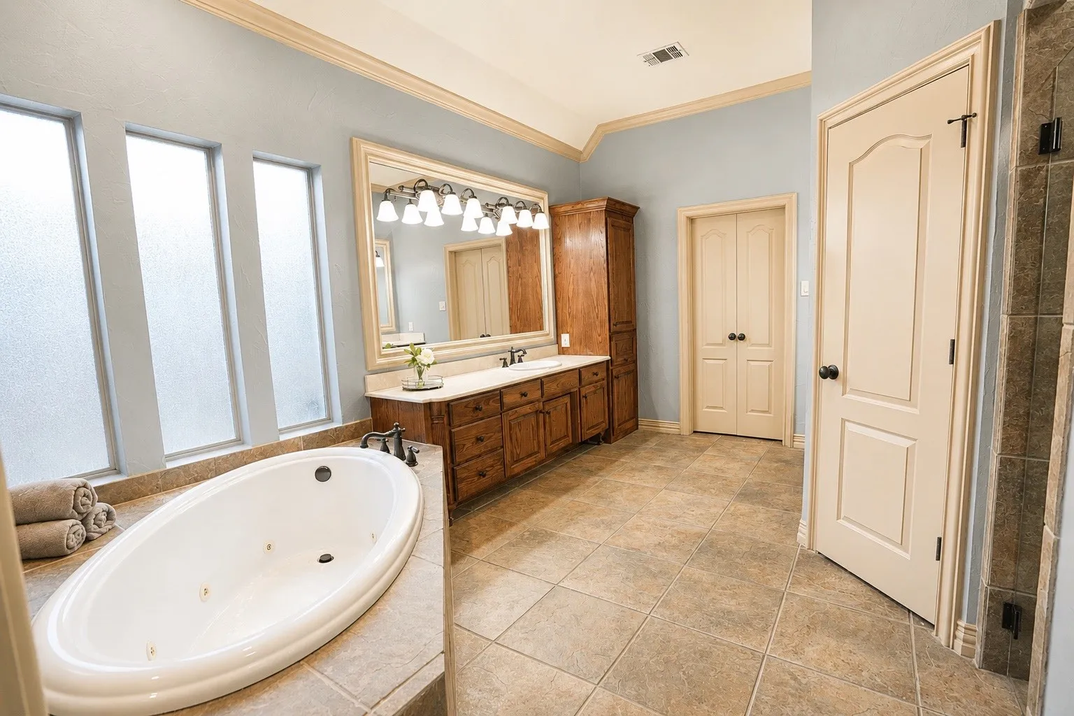 Soaking tub with tiled surround, wood-finish vanity with dual sinks, overhead lighting fixture, and tiled flooring
