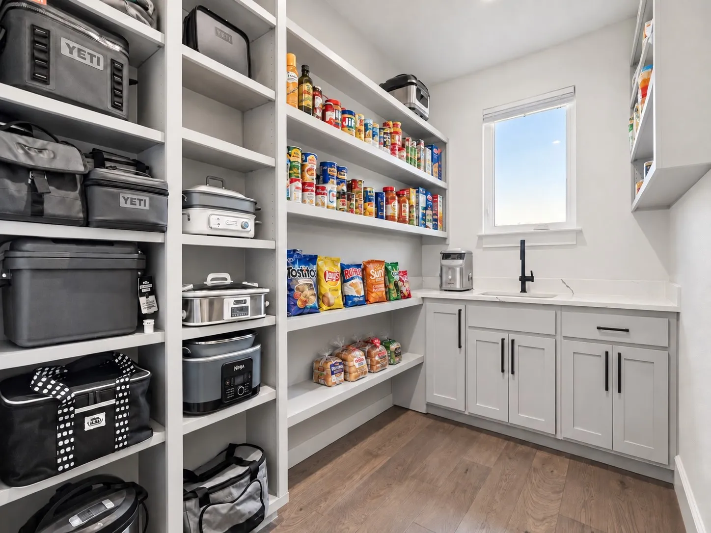 Spacious pantry featuring extensive built-in shelving, wood-finish flooring, white cabinetry with black hardware, a countertop, and an integrated sink with a black faucet