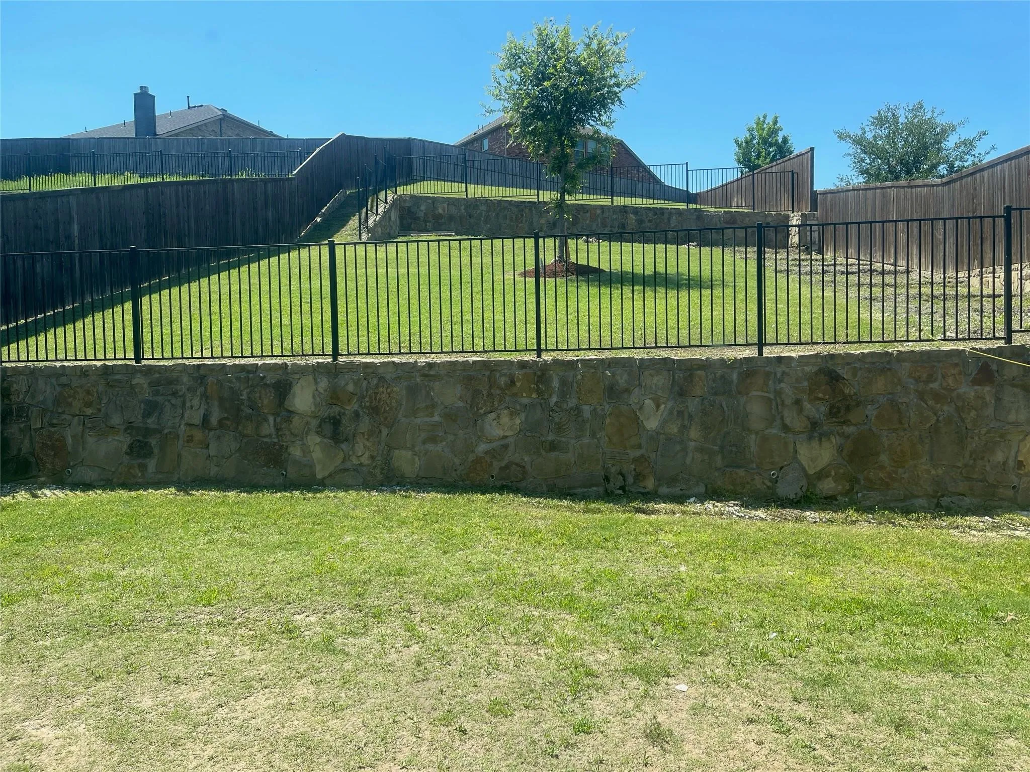 Tiered backyard featuring stone retaining walls, black metal fencing, and a manicured lawn