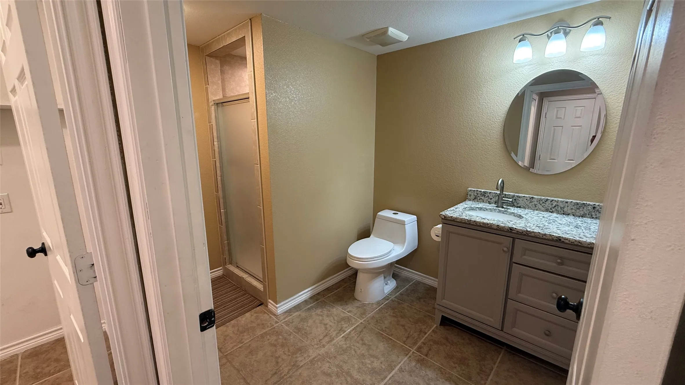 Bathroom featuring a gray vanity with a stone countertop, round mirror, and a shower with a frosted glass door