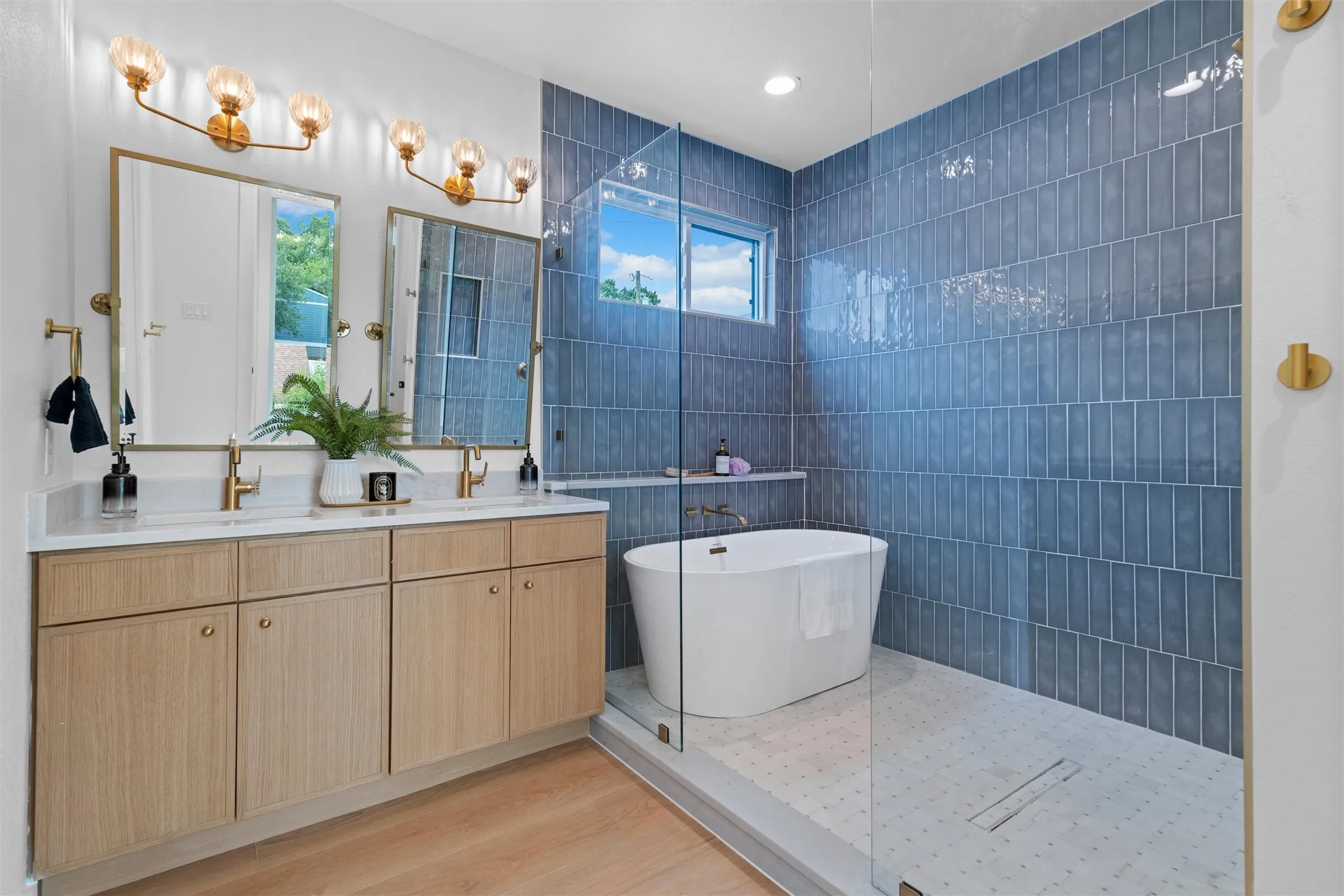 Bathroom featuring a freestanding soaking tub, a separate shower with blue vertical tile, a dual-sink vanity with light wood-finish cabinetry, and gold-finish fixtures
