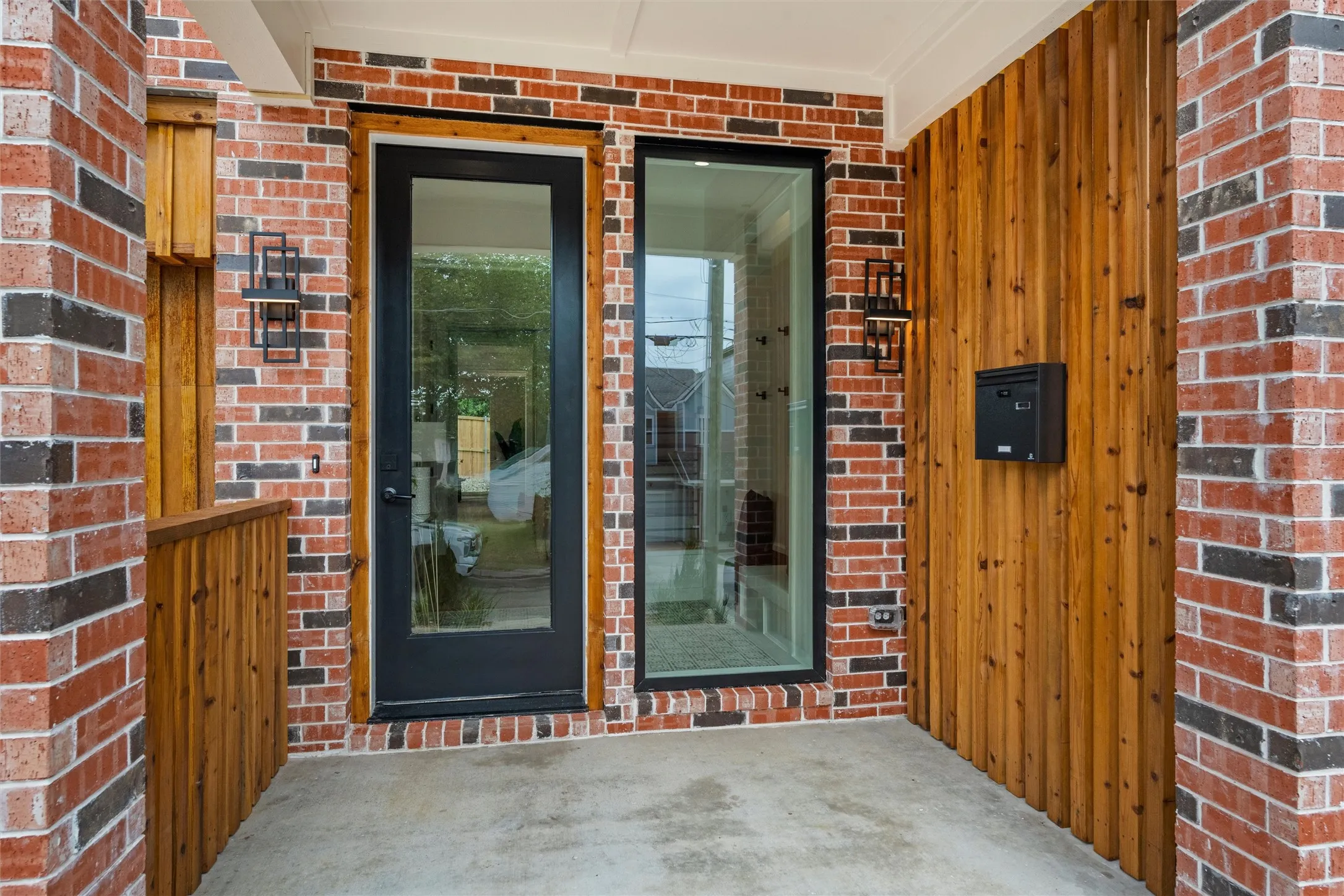 Entryway featuring brick and vertical wood siding, a full-lite door with a black frame, an accompanying full-length window, and modern exterior light fixtures
