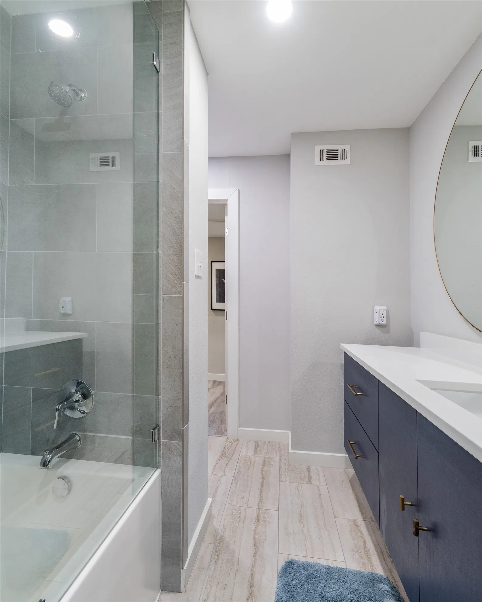 Integrated bathtub and shower with glass enclosure, light gray wall tile, white vanity countertop, navy blue vanity cabinetry, and wood-finish flooring