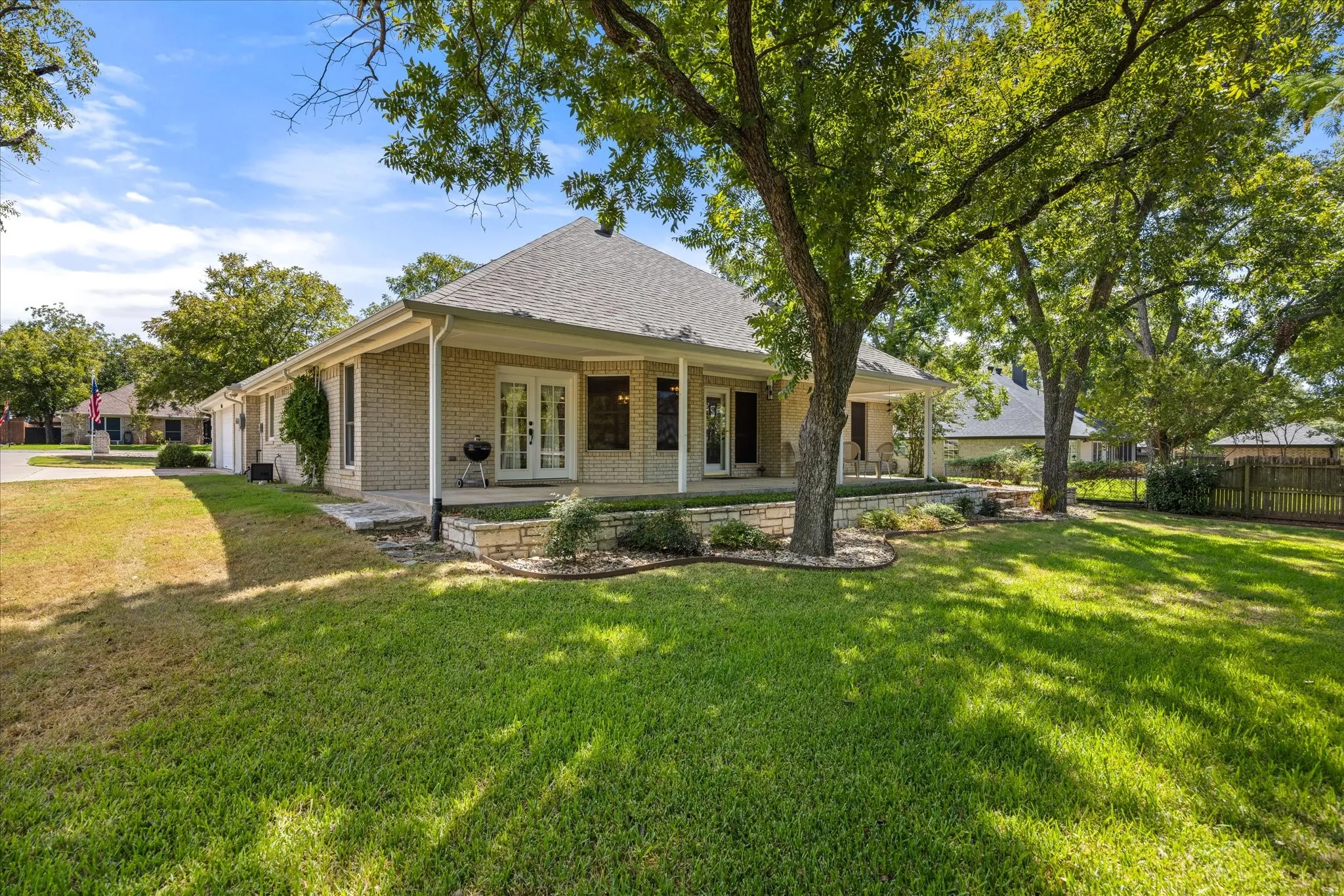 Expansive lawn with mature trees, brick exterior, covered patio, French doors, and stone landscaping accents