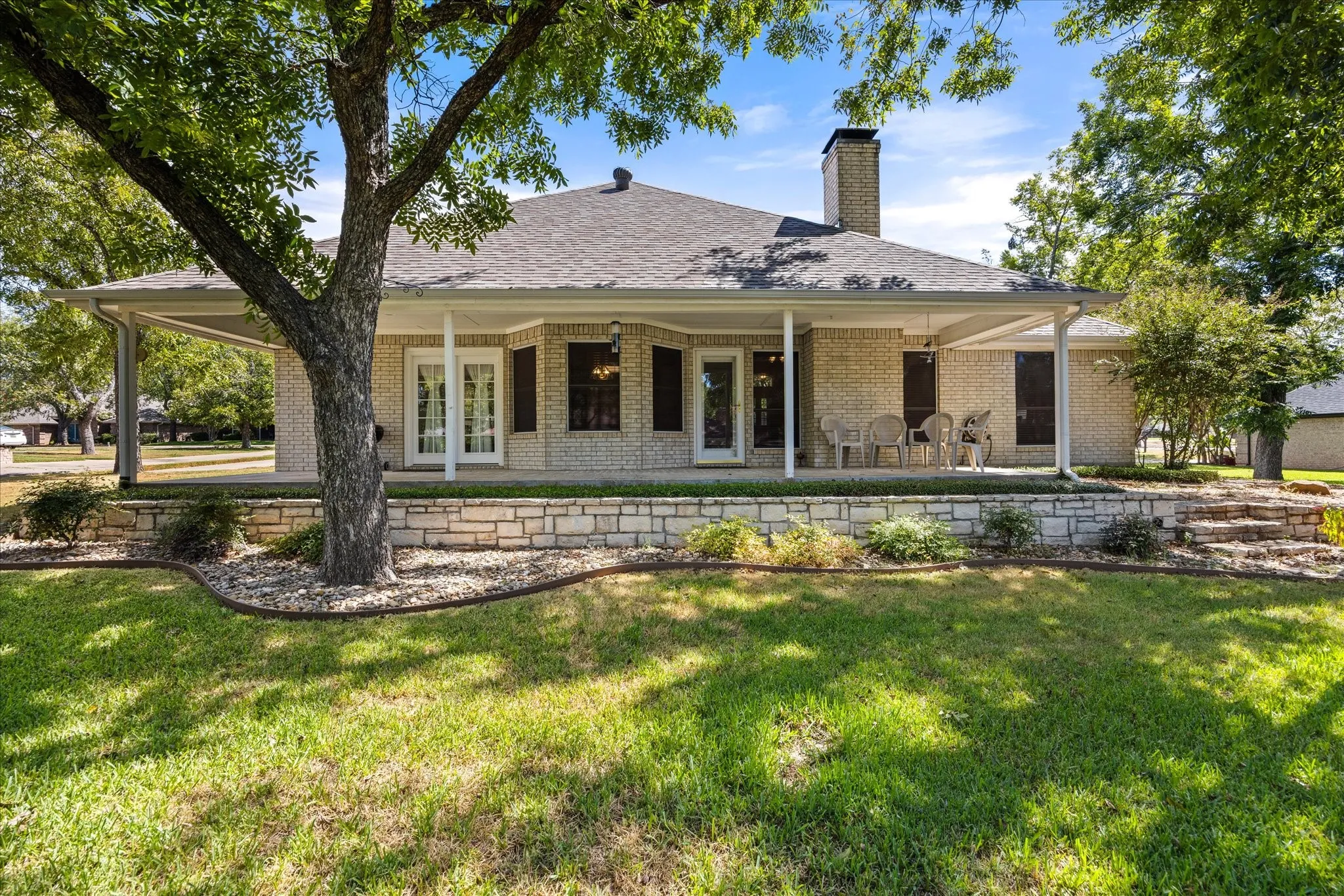 Brick exterior with a covered patio, stone retaining wall, and landscaped planting beds
