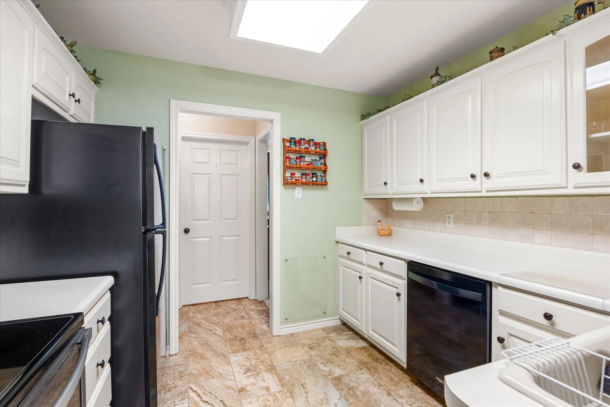 White cabinetry with dark hardware, stone-finish flooring, a neutral-toned tile backsplash, and a black built-in dishwasher
