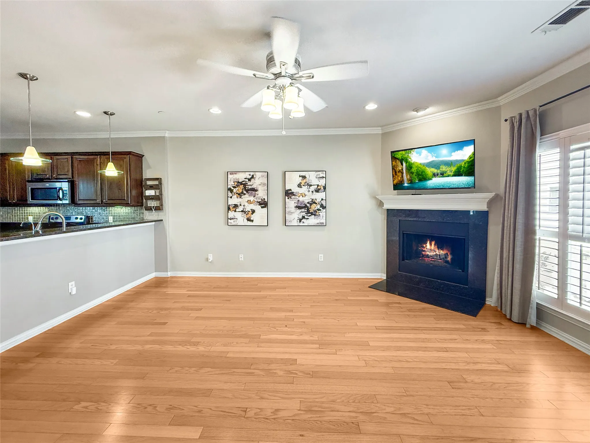 Wood-finish flooring throughout, ceiling fan with integrated lighting, built-in fireplace with dark stone surround, white window shutters, and recessed lighting