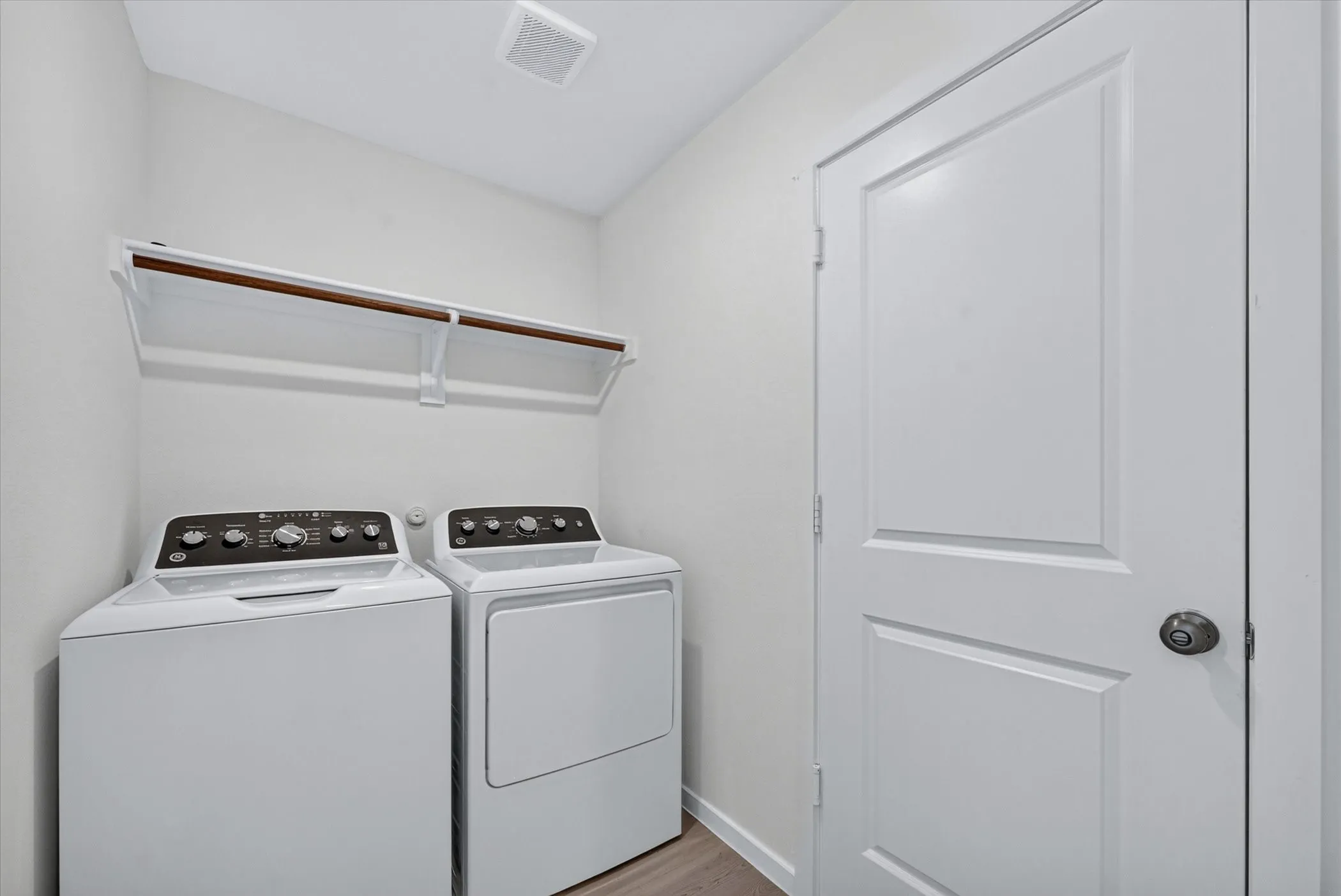 Dedicated laundry space featuring a white top-load washer and dryer, an overhead storage shelf with a hanging rod, and light wood-finish flooring