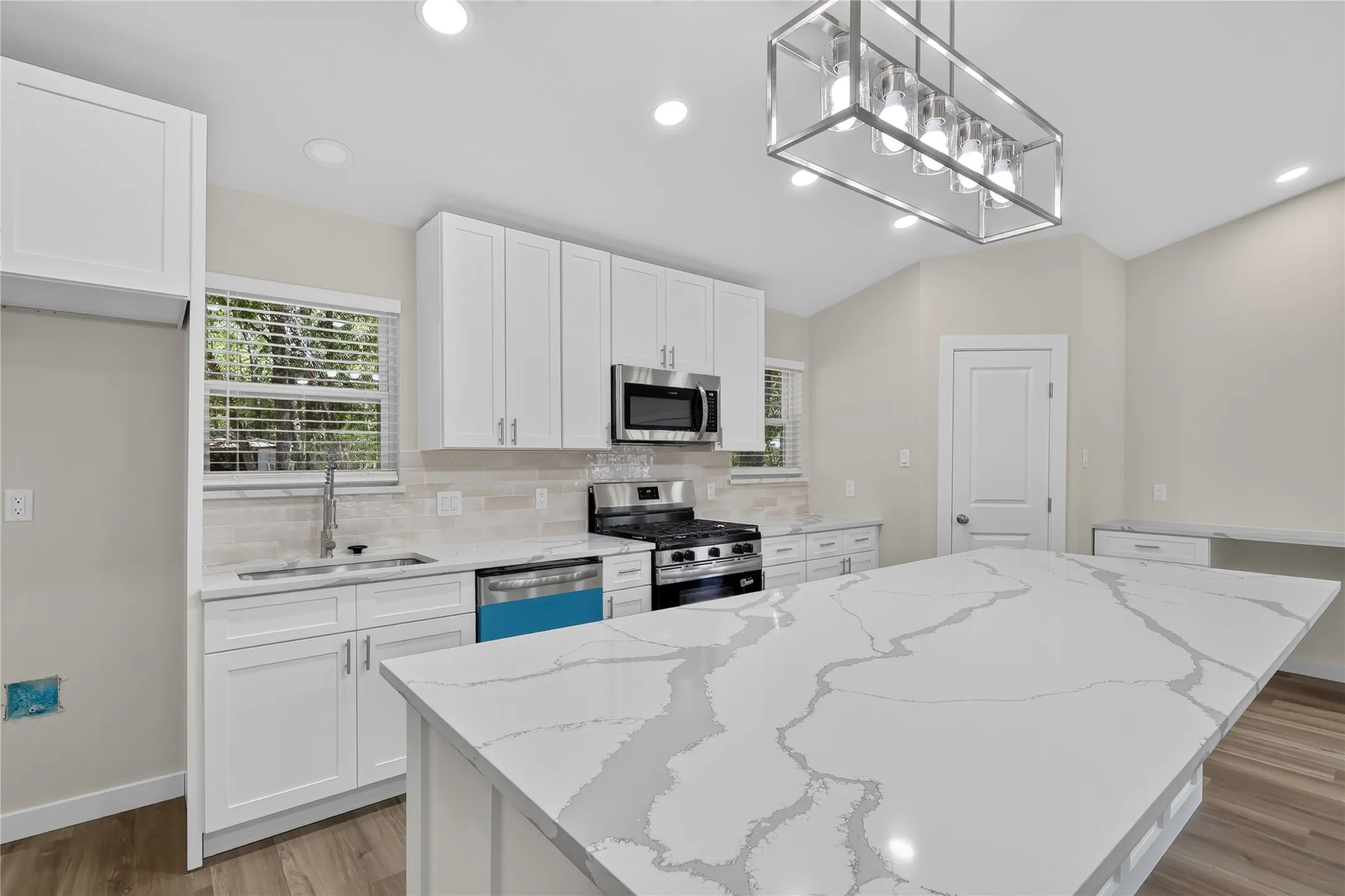 Spacious kitchen featuring an expansive island, white shaker cabinetry, stainless steel appliances, modern pendant lighting, and wood-finish flooring
