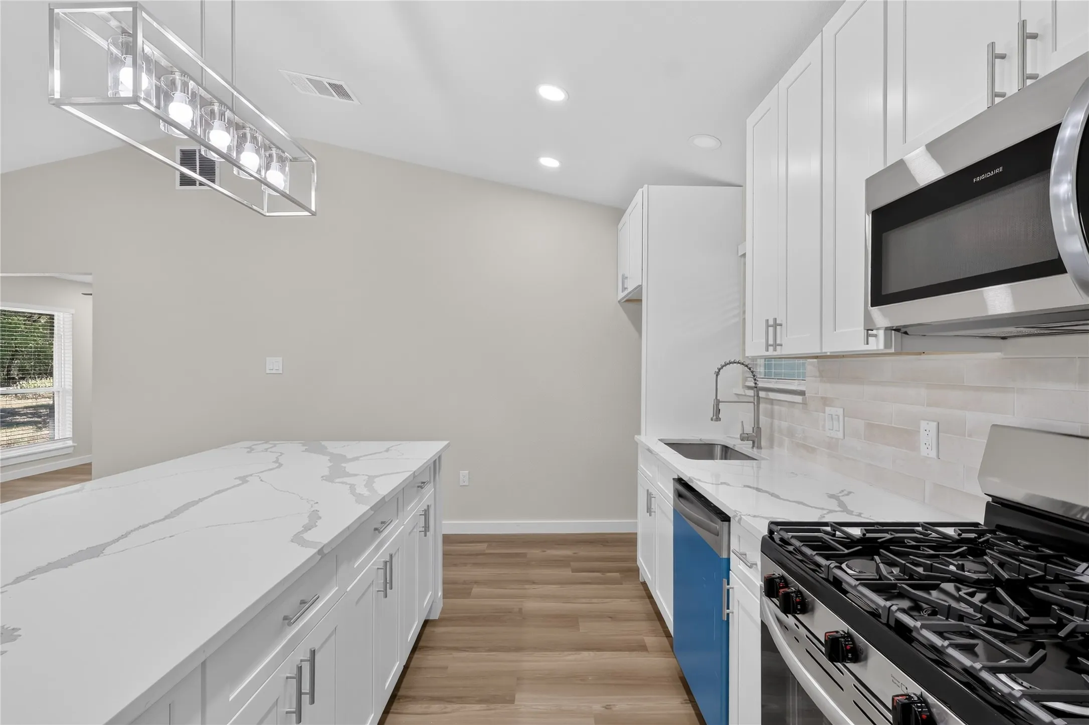 Vaulted ceiling with recessed lighting, wood-finish flooring, white cabinetry with silver-tone hardware, large kitchen island with a quartz countertop, and stainless steel appliances