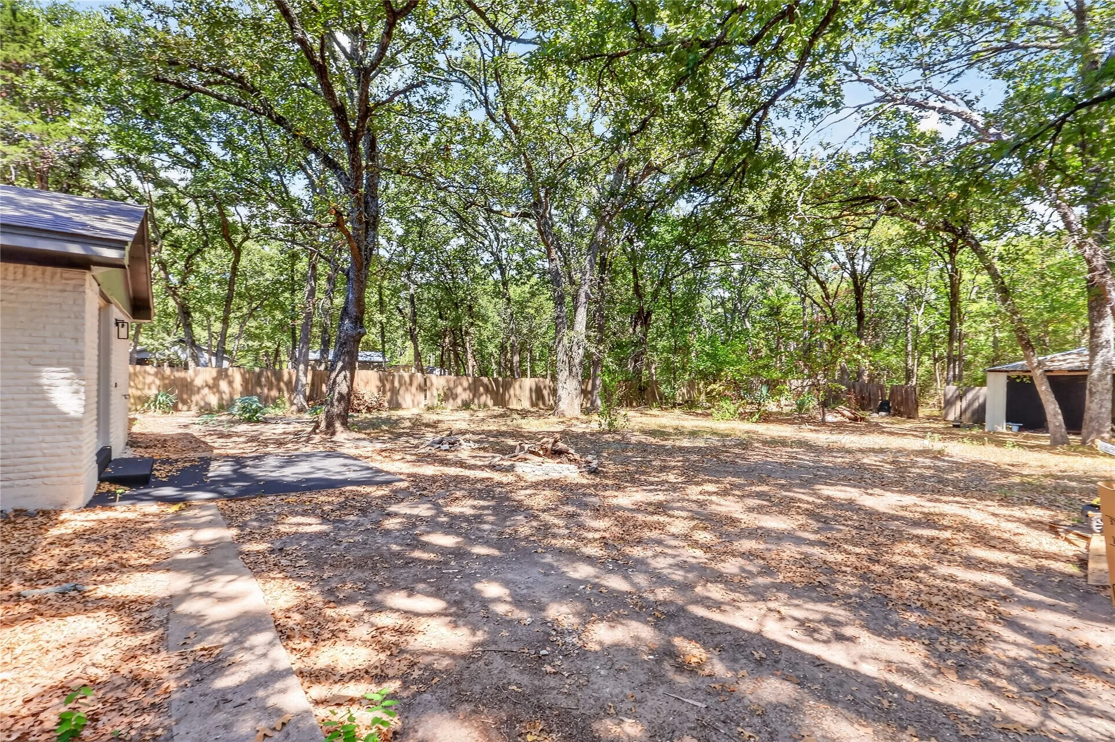 Expansive outdoor area featuring mature trees, a wooden fence perimeter, a detached auxiliary structure, a stone pathway, and a patio area