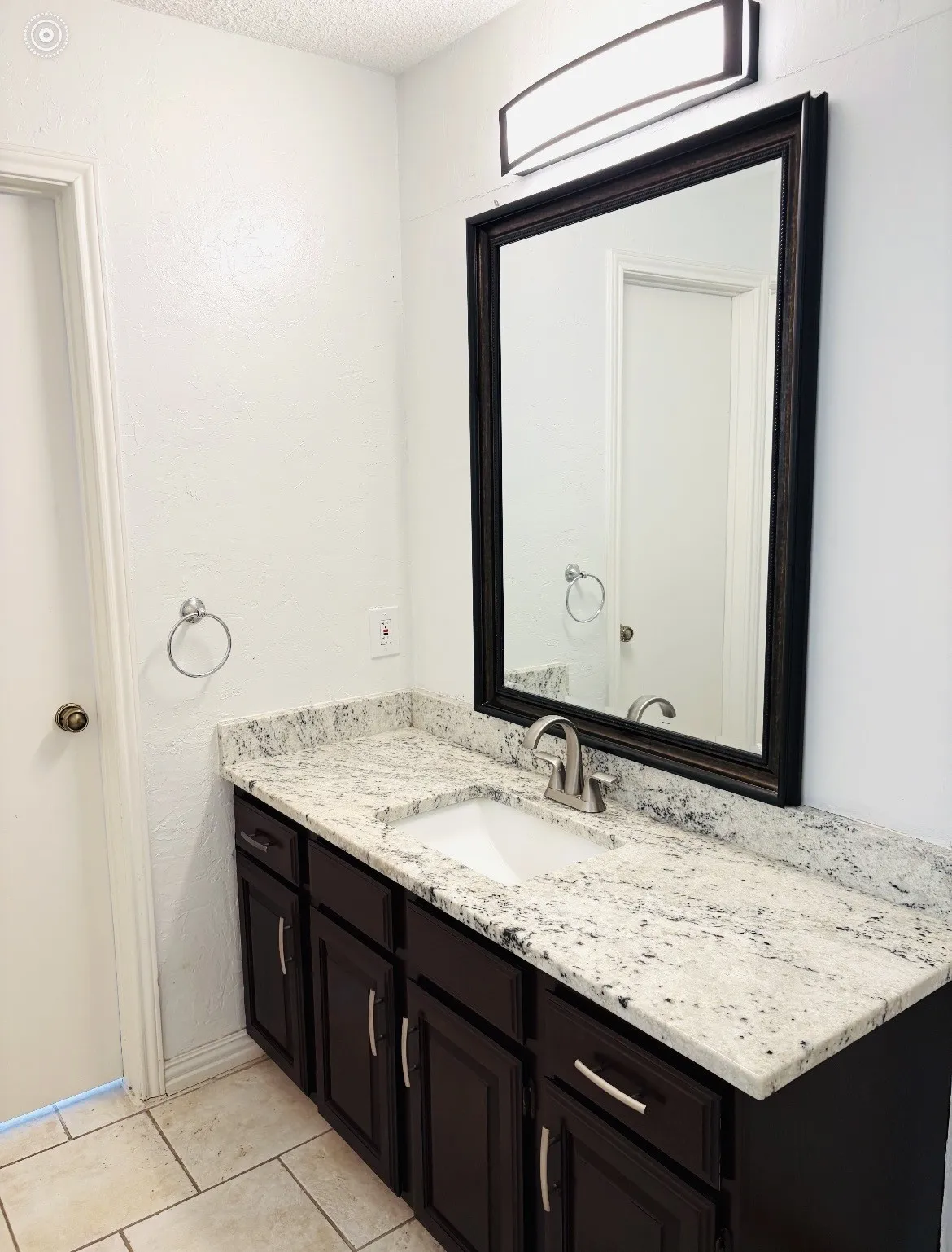 White countertop with integrated sink and chrome faucet, dark wood-finish vanity with chrome pulls, framed mirror, and rectangular wall sconce