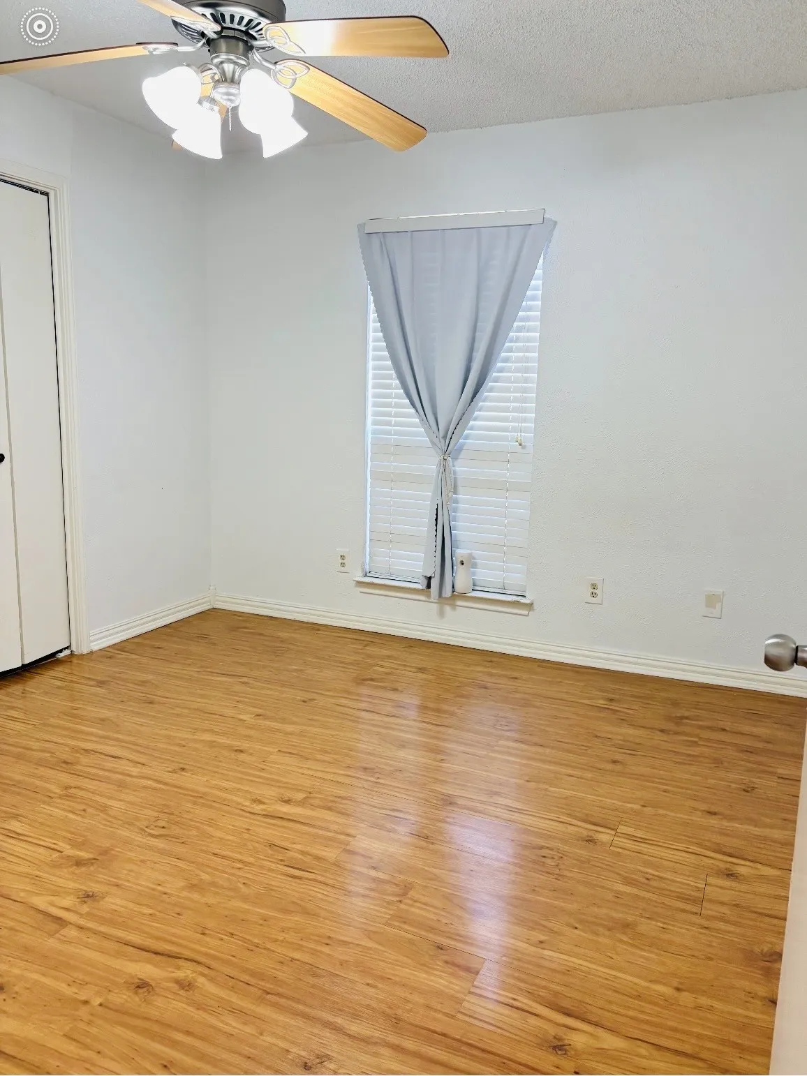 Wood-finish flooring throughout, bright white walls, window with blinds, and ceiling fan with integrated lighting