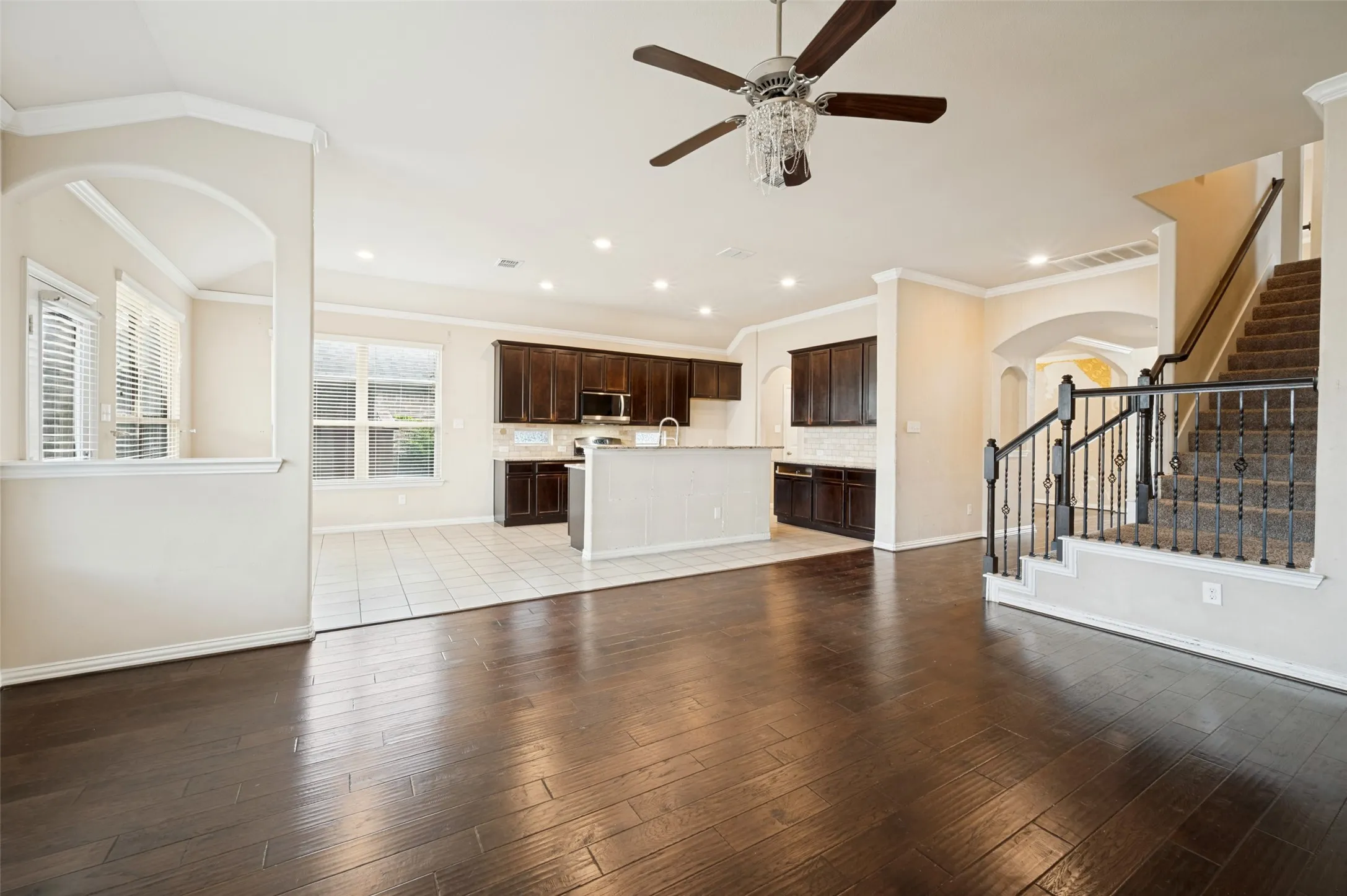 Dark wood-finish flooring, recessed lighting, ceiling fan with chandelier lighting, dark wood cabinetry, and a carpeted staircase with an ornamental wrought-iron railing