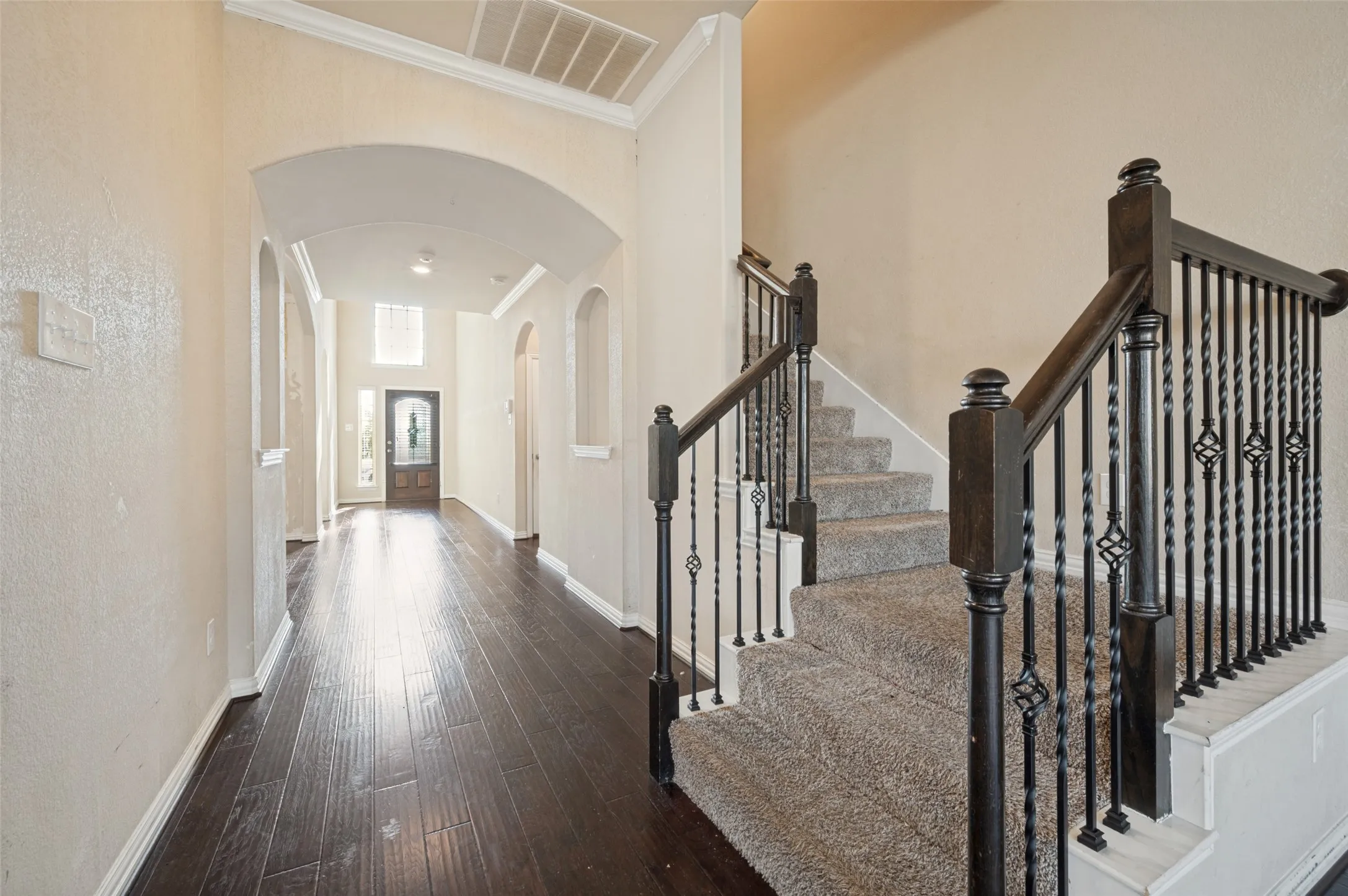 Dark wood-finish flooring, staircase with wrought iron balusters, arched doorways, crown molding, and a paneled front door with decorative glass