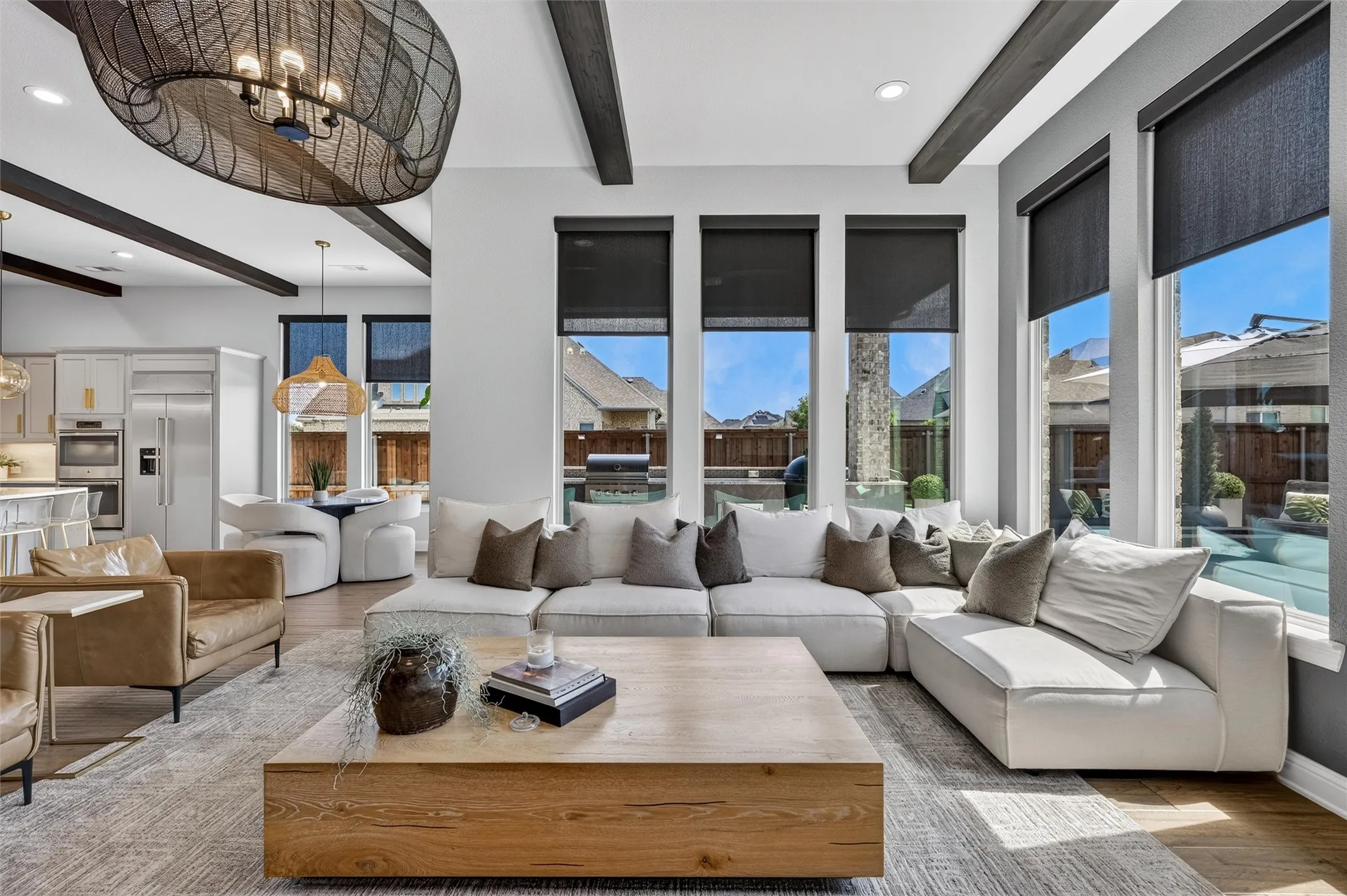 Open-concept living area with exposed wood beams, a prominent chandelier, and multiple tall windows featuring roller shades