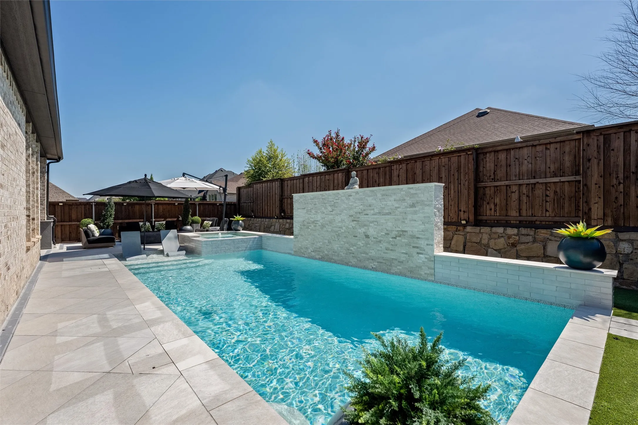 Outdoor entertaining space featuring a rectangular swimming pool, integrated spa, tiled water feature, and extensive light-toned stone-finish patio
