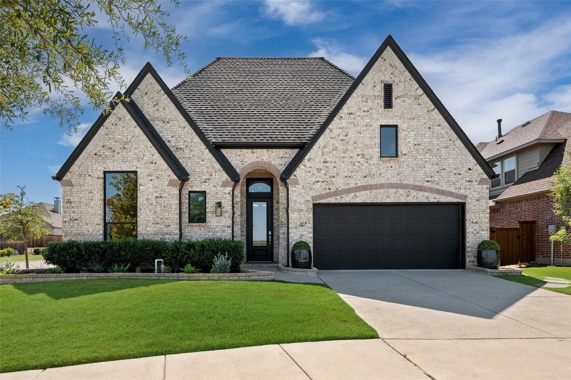 Light-colored brick exterior with dark trim accents, multiple gables, a two-car garage with a dark door, arched entry, and a manicured front lawn