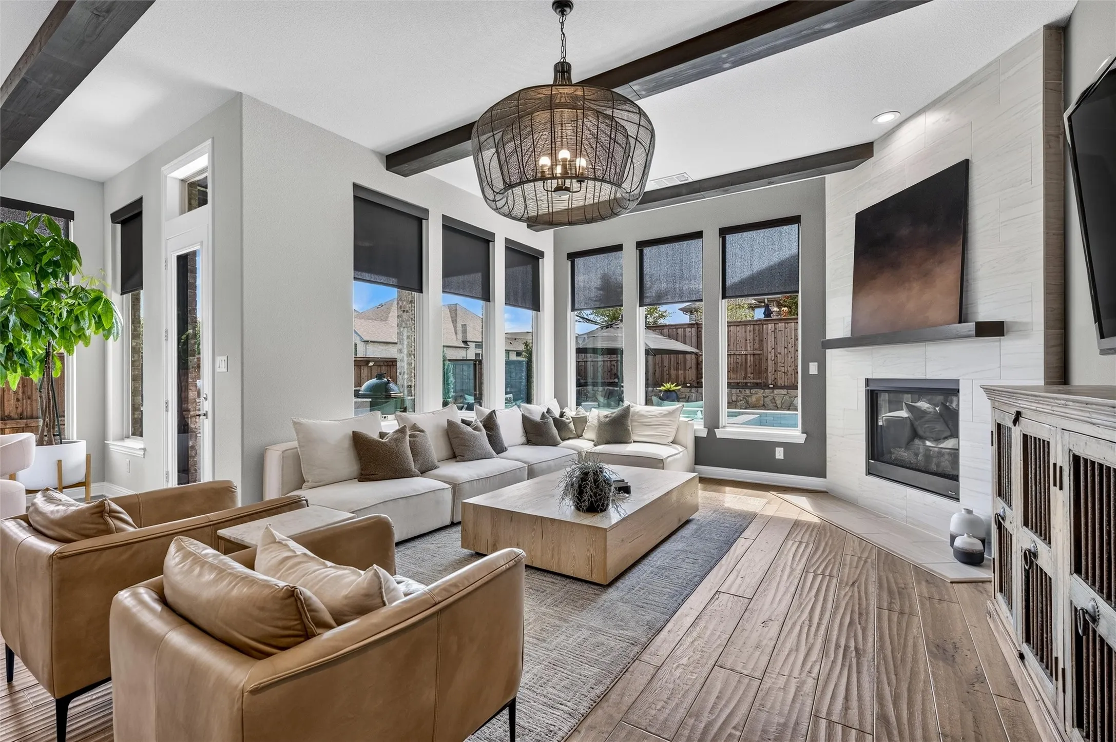 Spacious living area featuring exposed ceiling beams, a modern fireplace with a tiled surround, expansive windows with roller shades, and wood-finish flooring